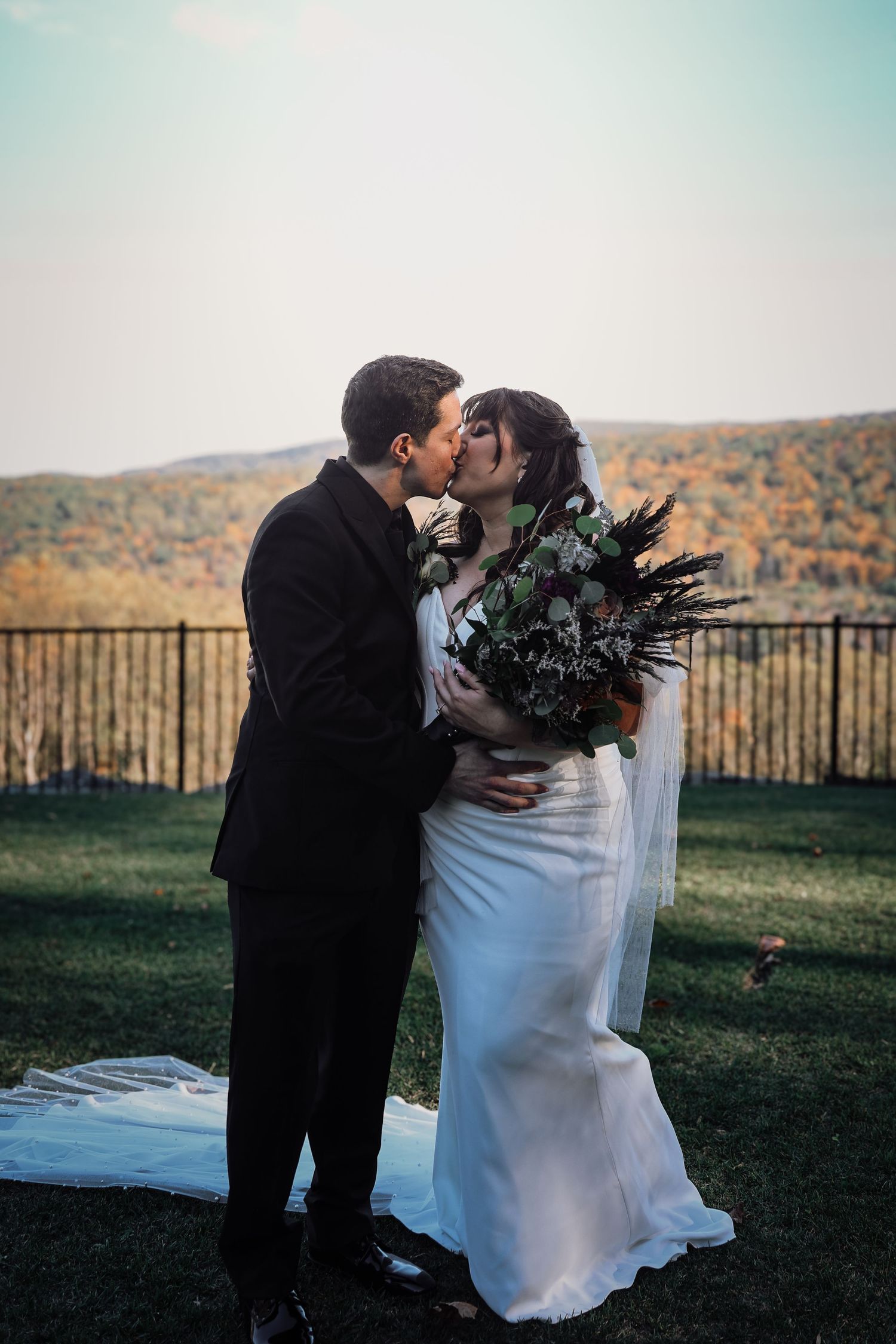 A romantic wedding couple shares a tender kiss against a backdrop of autumn mountains and rolling hills at sunset.