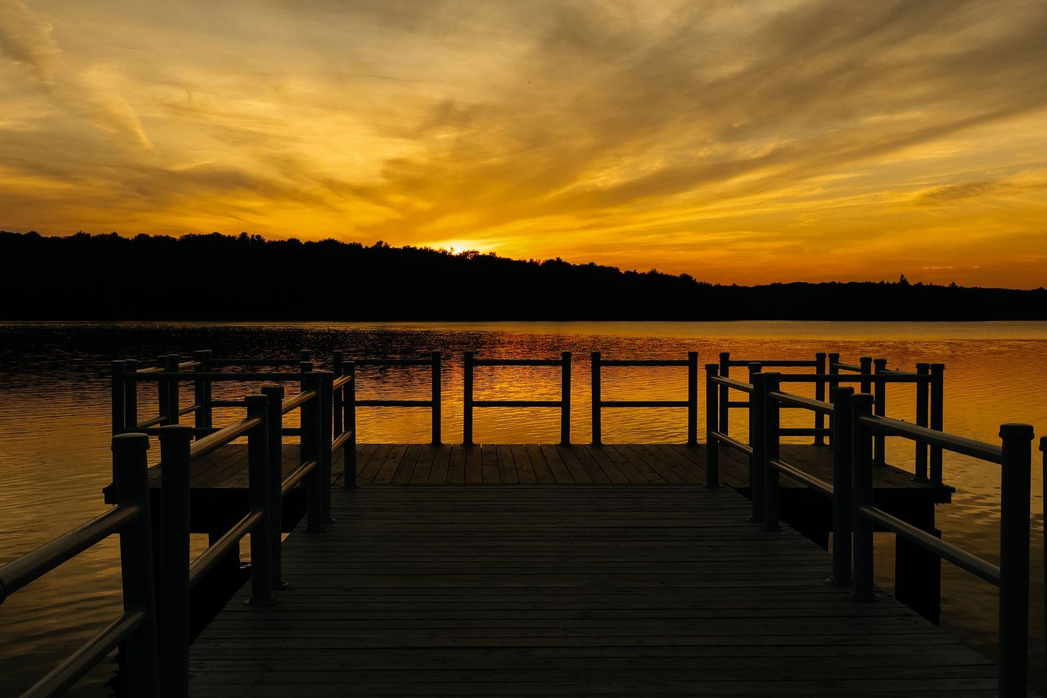 Wooden dock on calm lake during dramatic golden sunset with orange sky and silhouetted treeline in background.