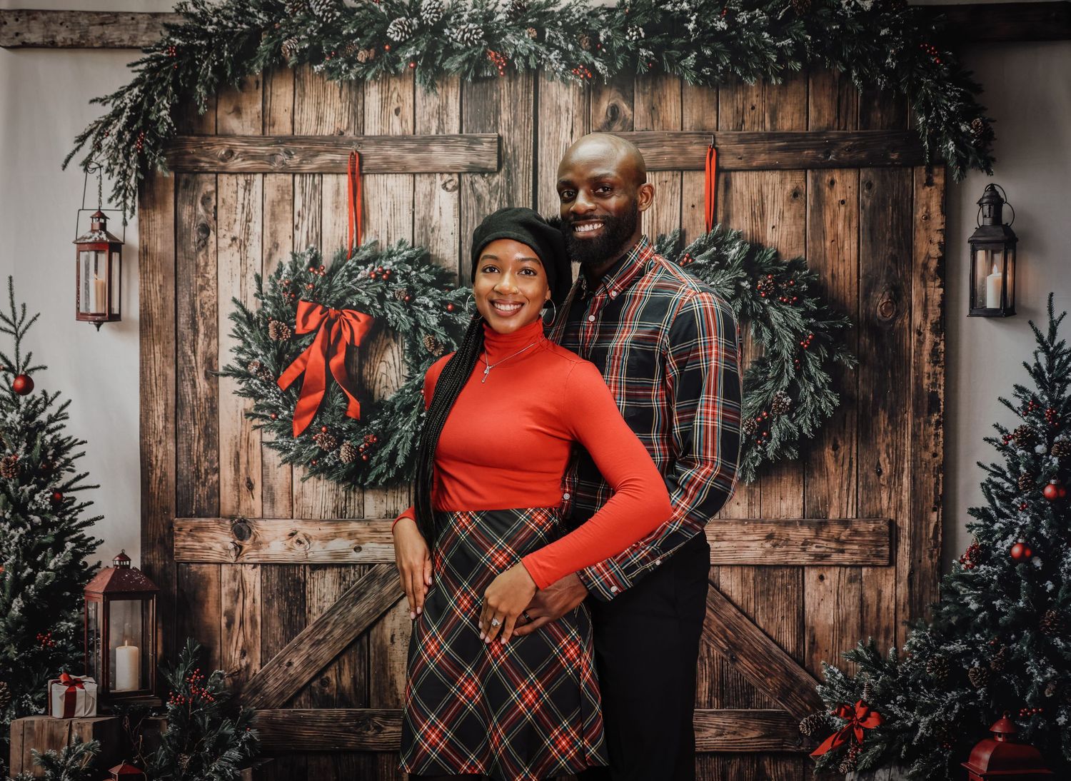 Holiday portraits with plaid dress and red sweater against rustic wooden door with evergreen garland decorations.