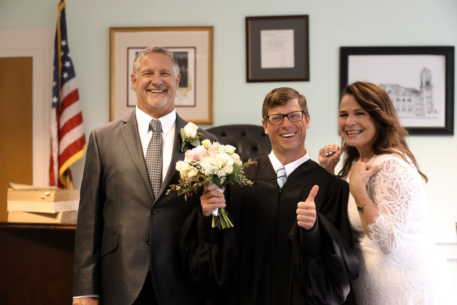 Three people smile together in a courthouse with an American flag and framed artwork on the wall, one holding white flowers.