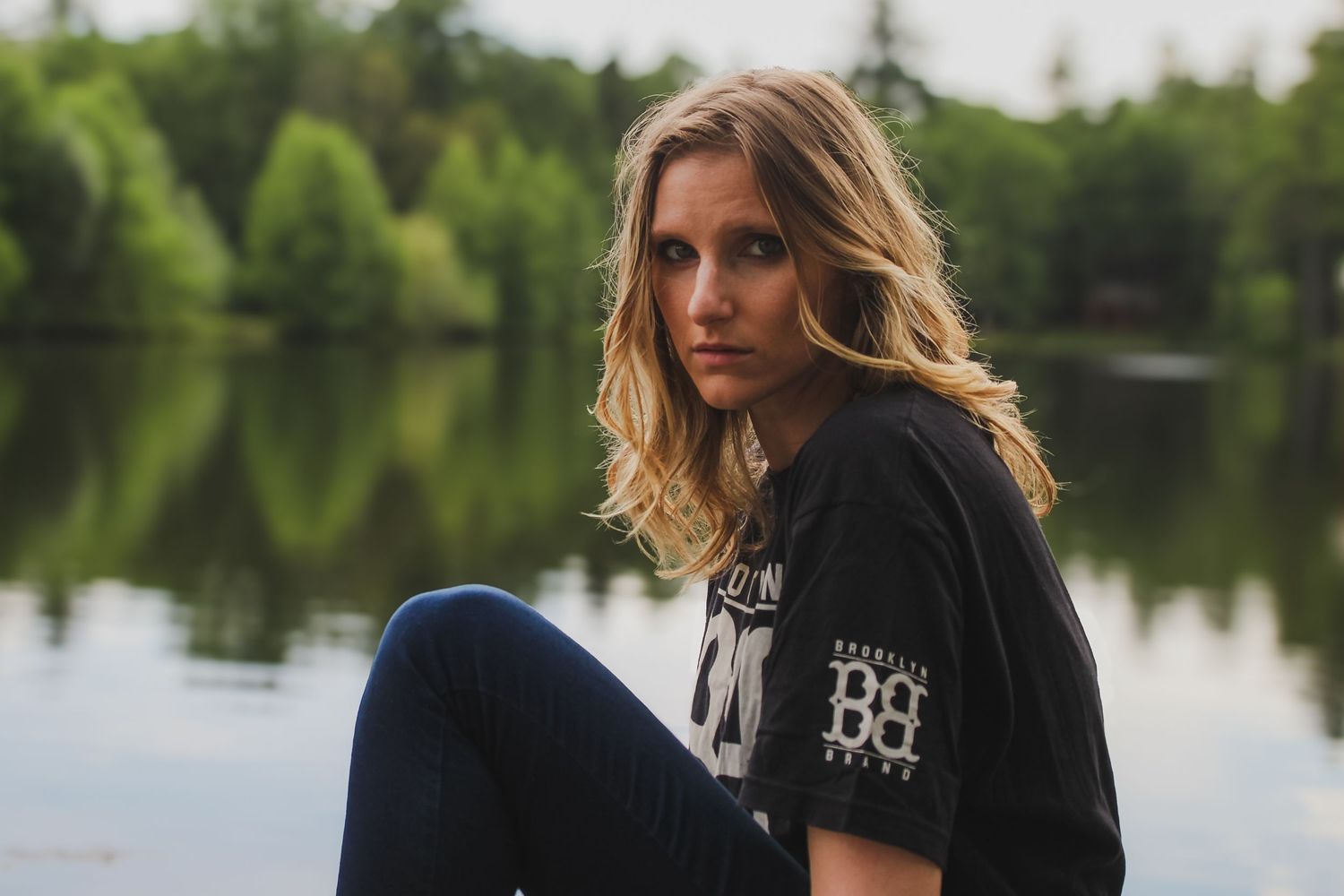 Person in a black t-shirt sits by a serene lake surrounded by green trees on a calm day.