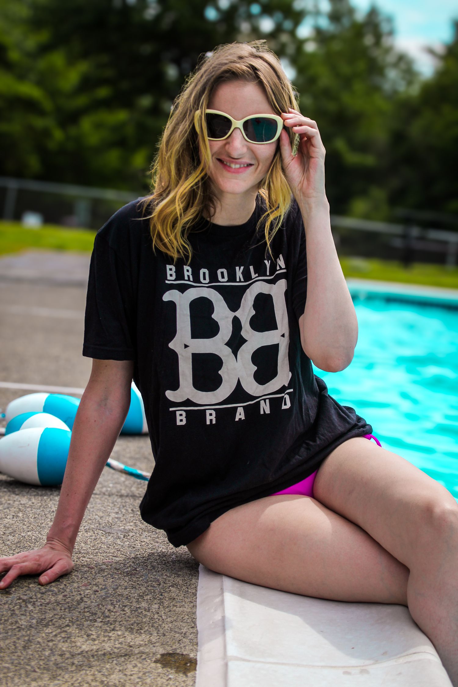 Woman in black Brooklyn Brand t-shirt and sunglasses relaxing by swimming pool on sunny summer day.