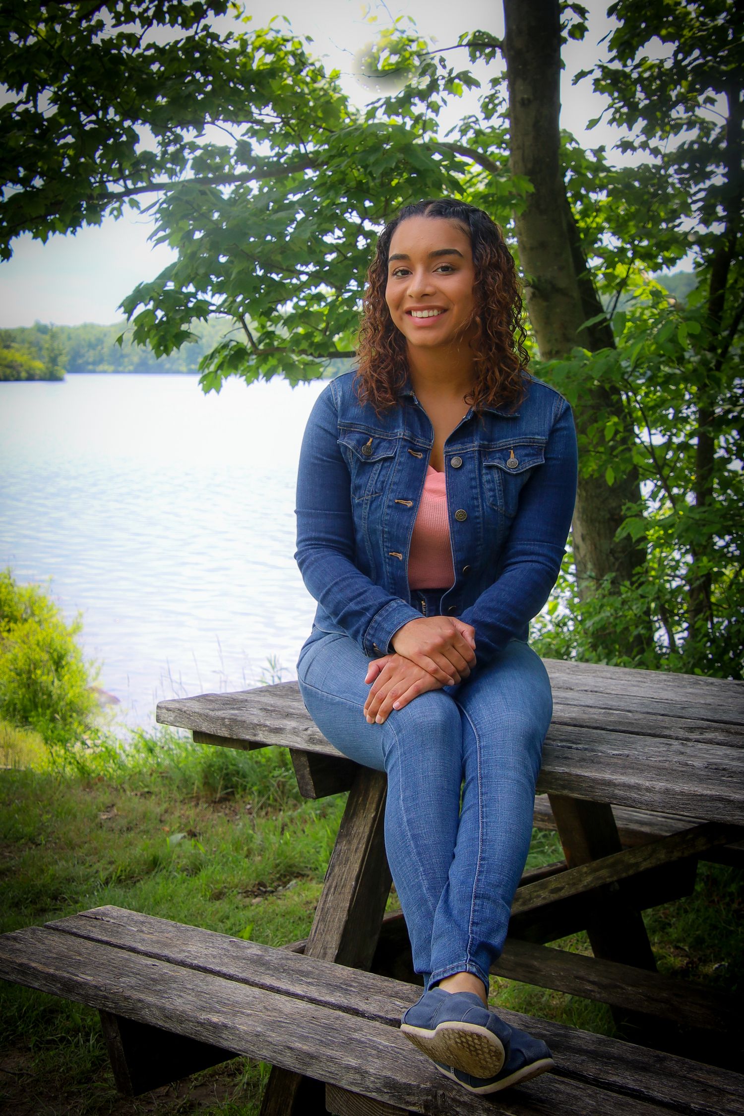 Someone in denim sits casually on a weathered wooden bench by a serene lakeside surrounded by lush summer foliage.