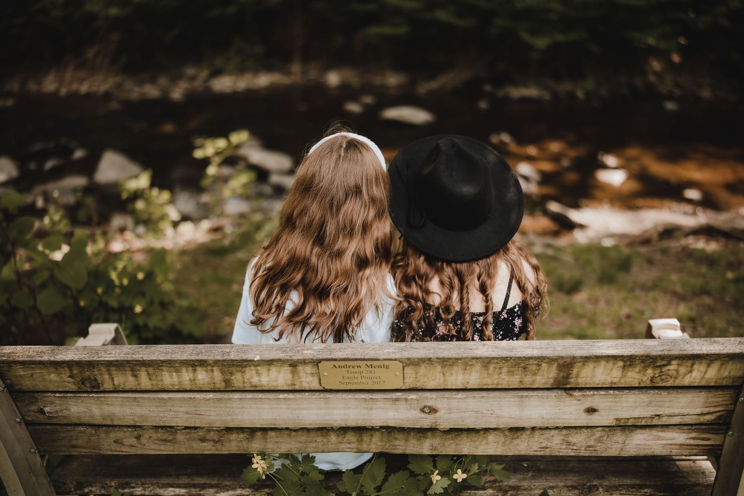 Two people with long hair wearing black sit together on a wooden bench in an outdoor garden setting.
