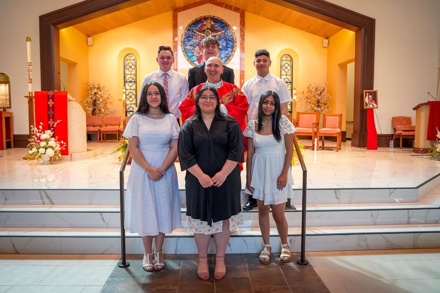 People gather in a church sanctuary with stained glass windows and altar area illuminated by warm lighting.