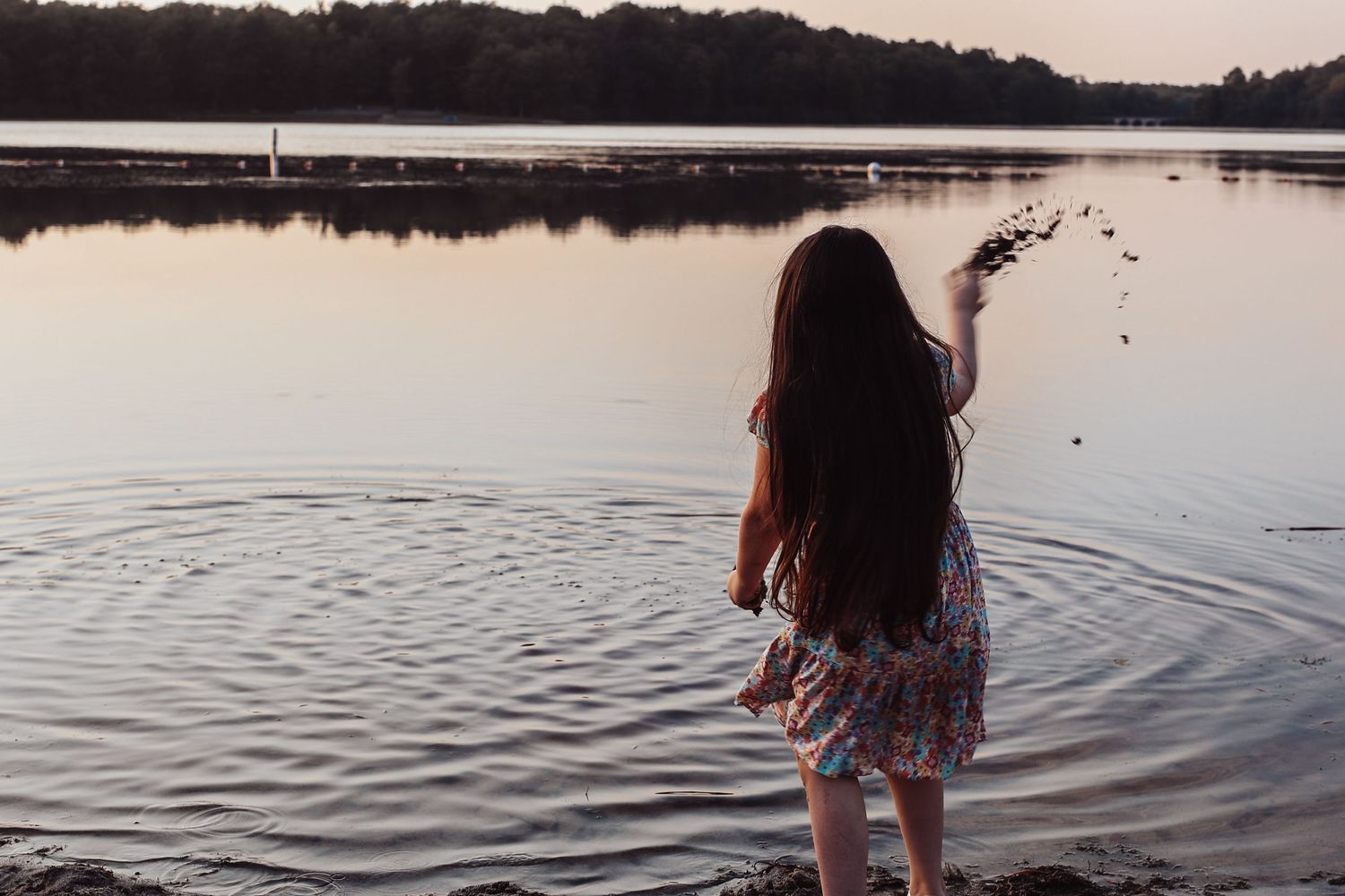 A person stands at the edge of a tranquil lake during sunset, creating ripples in the calm water.