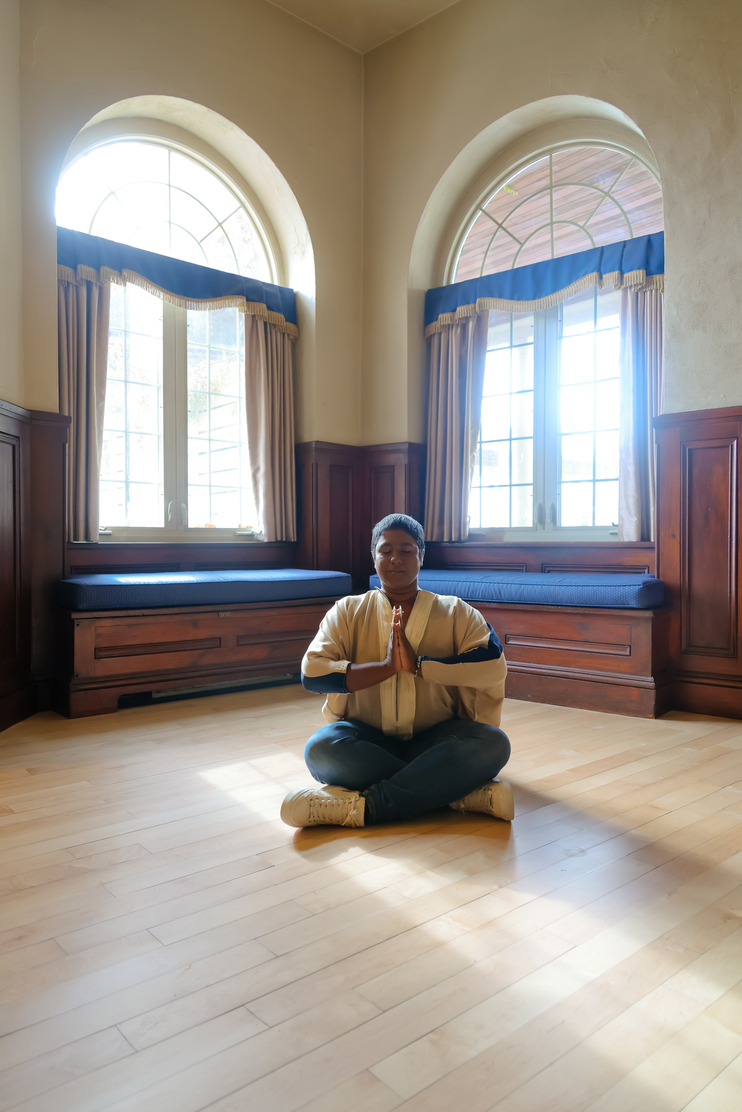 A figure sits in a sunlit room with arched windows and hardwood floors, reading while positioned in different yoga poses.