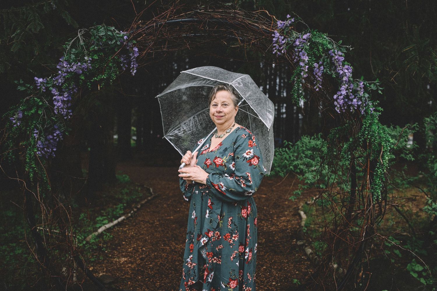 A person poses with an umbrella in a lush forest setting wearing a floral dress on a rainy day.