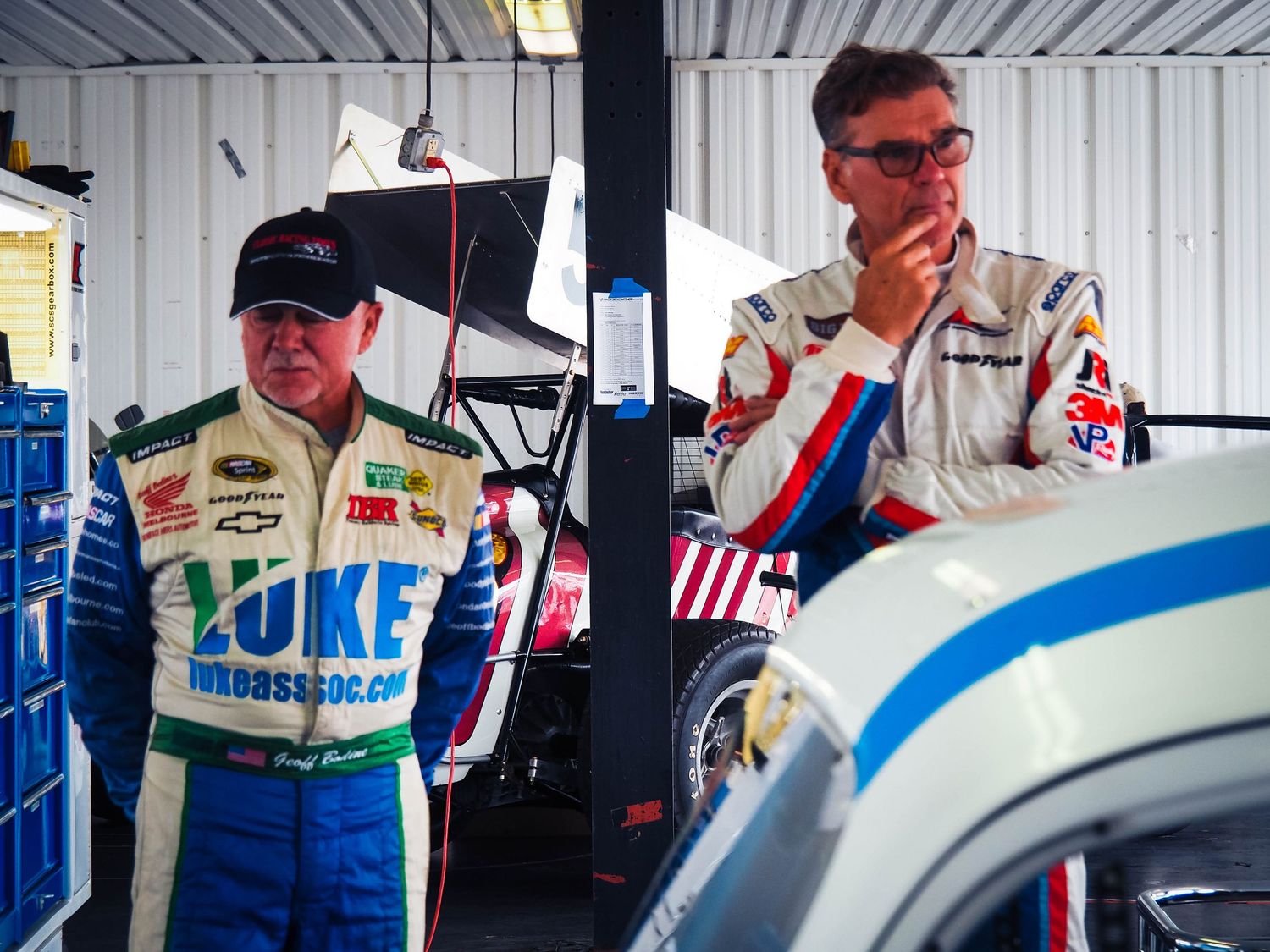 Race car drivers in racing suits stand near their vehicle in a garage during a motorsport event.