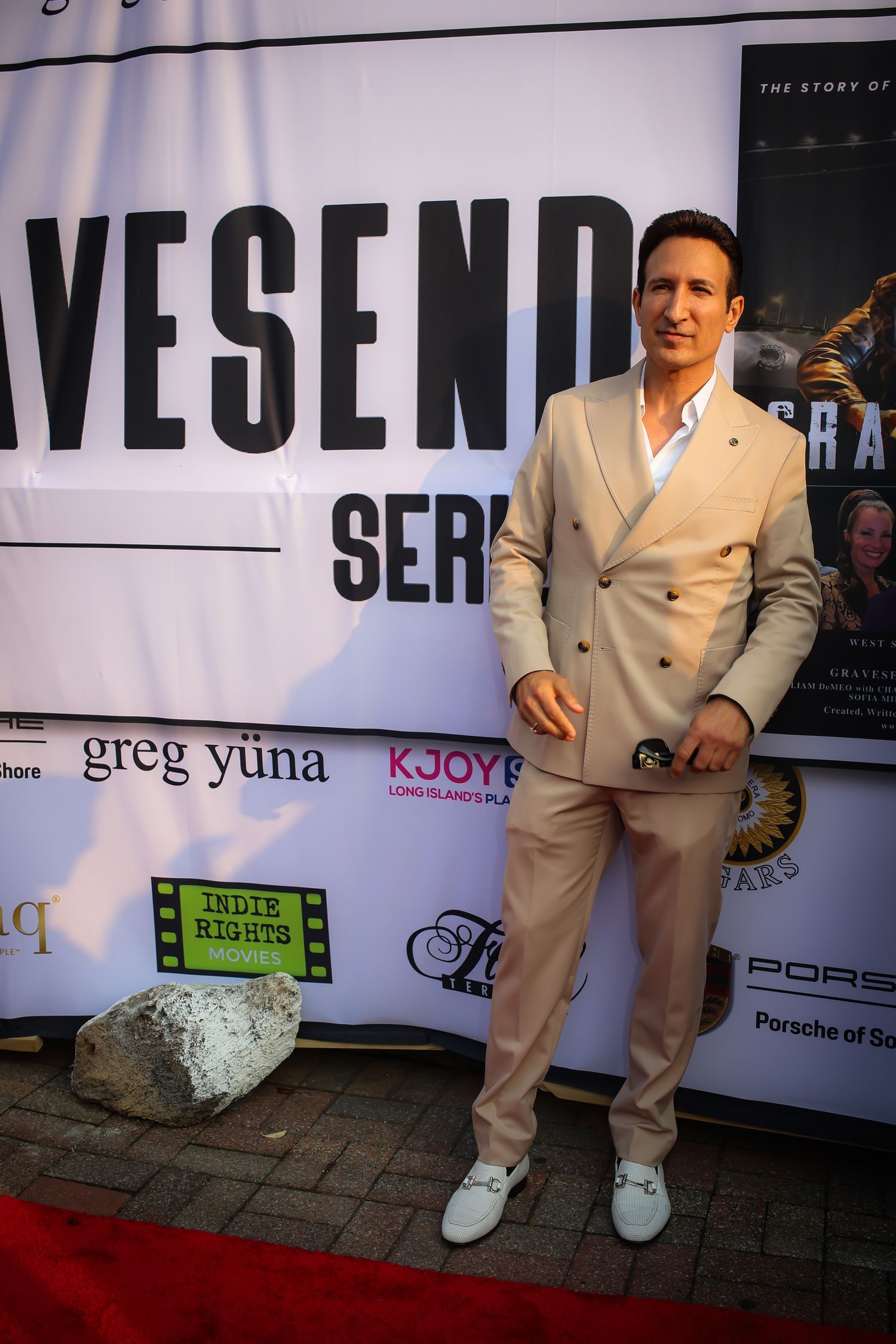 A person in a light-colored suit stands on a red carpet at an event with promotional banners and signage in the background.