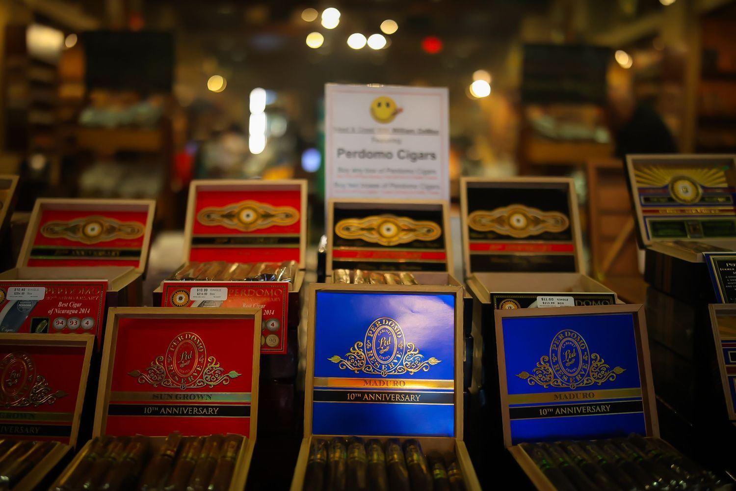 Rows of premium cigar boxes in red and blue colors displayed at a tobacco shop with soft bokeh lighting in background.