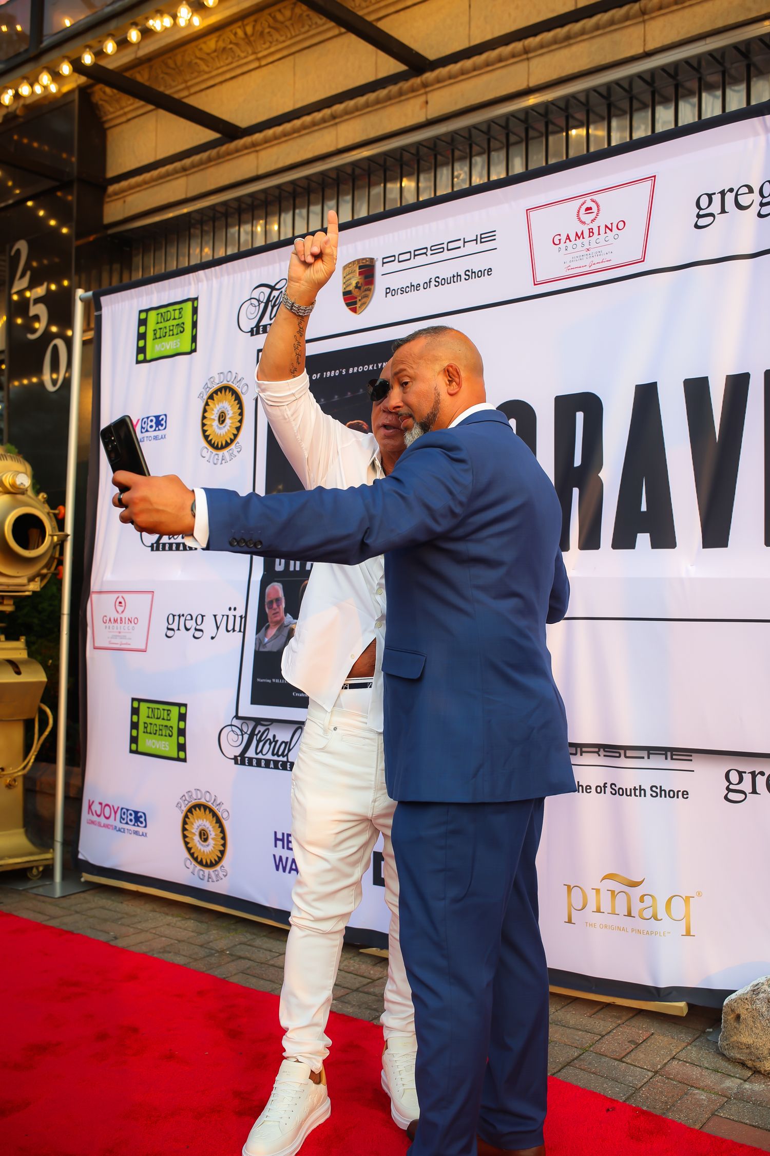 Two people in contrasting outfits pose together on a red carpet at an event with sponsor banners in the background.