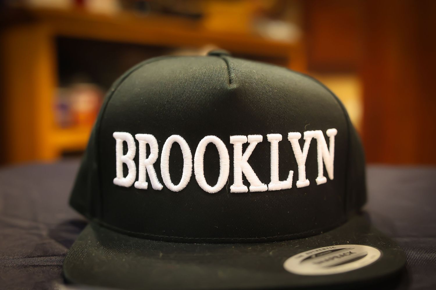 Black snapback baseball cap with Brooklyn text embroidered in white displayed on dark surface.