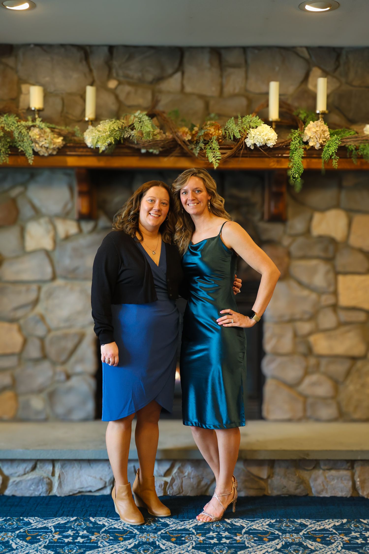 Two women in blue dresses standing on patterned carpet in front of stone wall with decorative lighting and greenery.