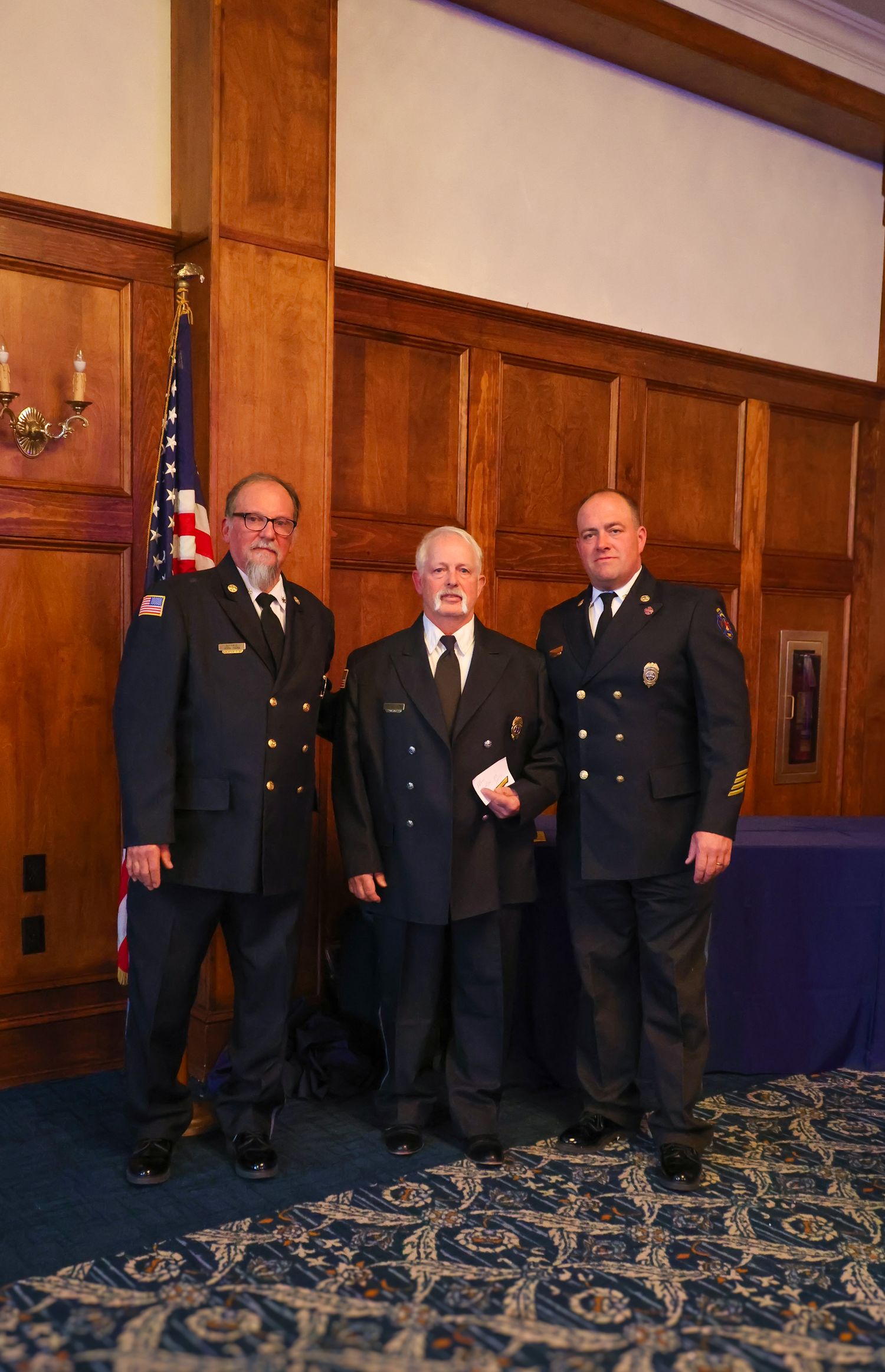 Three uniformed officers stand together in front of wood-paneled wall with American flag in formal ceremony setting.