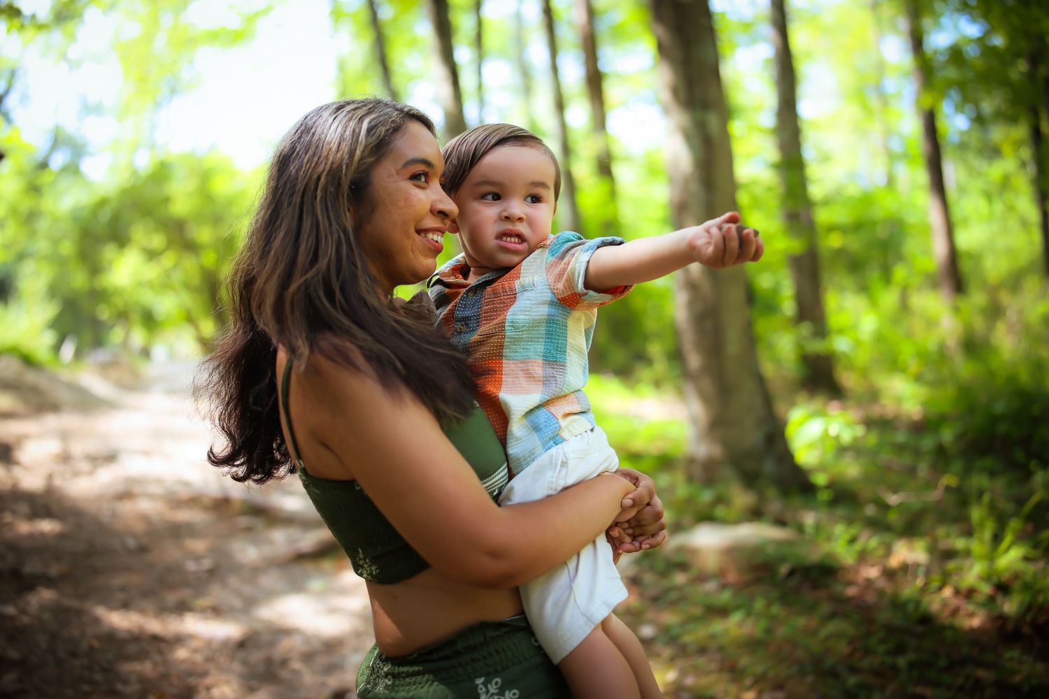 A parent and child share a joyful moment pointing at something while exploring a sunlit forest path.
