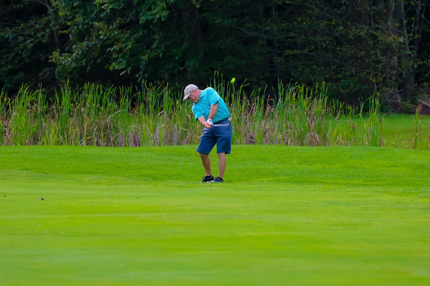 Golfer takes a swing on a lush green golf course with tall grass and trees in the background.