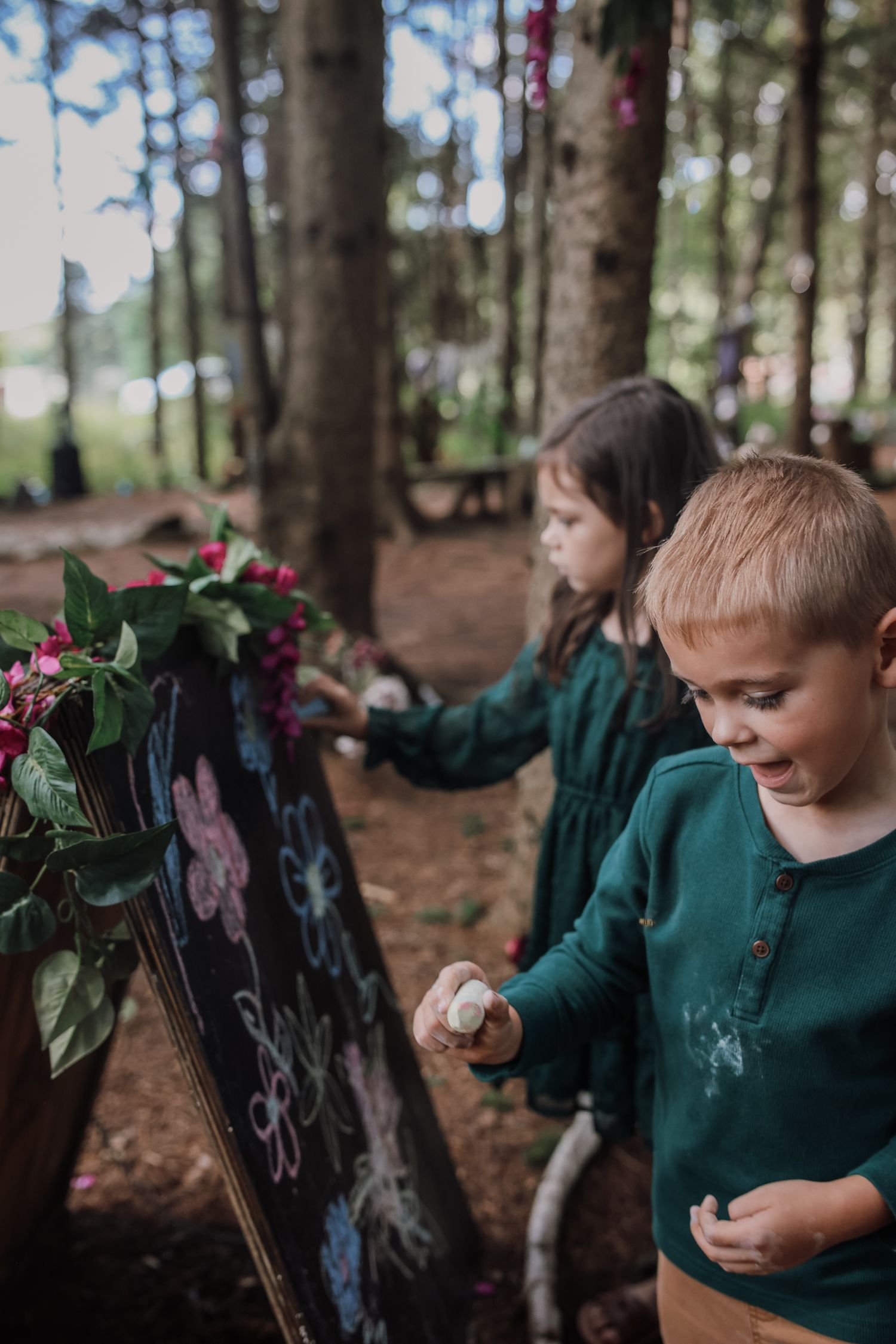 Children in green sweaters explore a wooded forest area with scattered trees and natural elements.