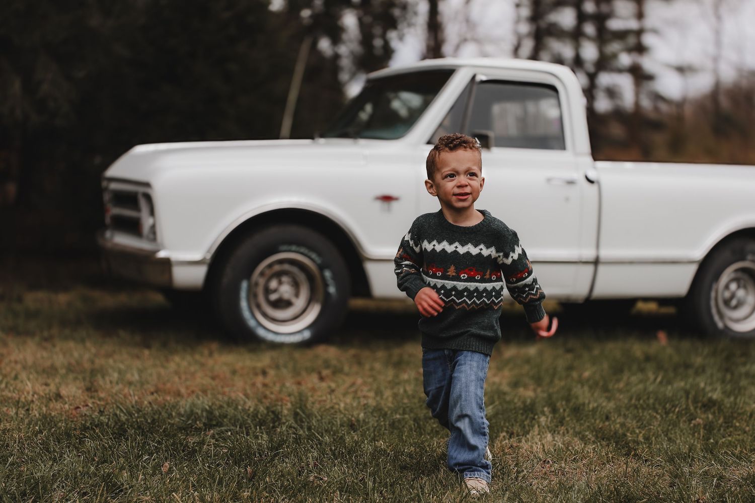 A person in a patterned sweater and jeans runs playfully through a grassy field on an overcast autumn day.