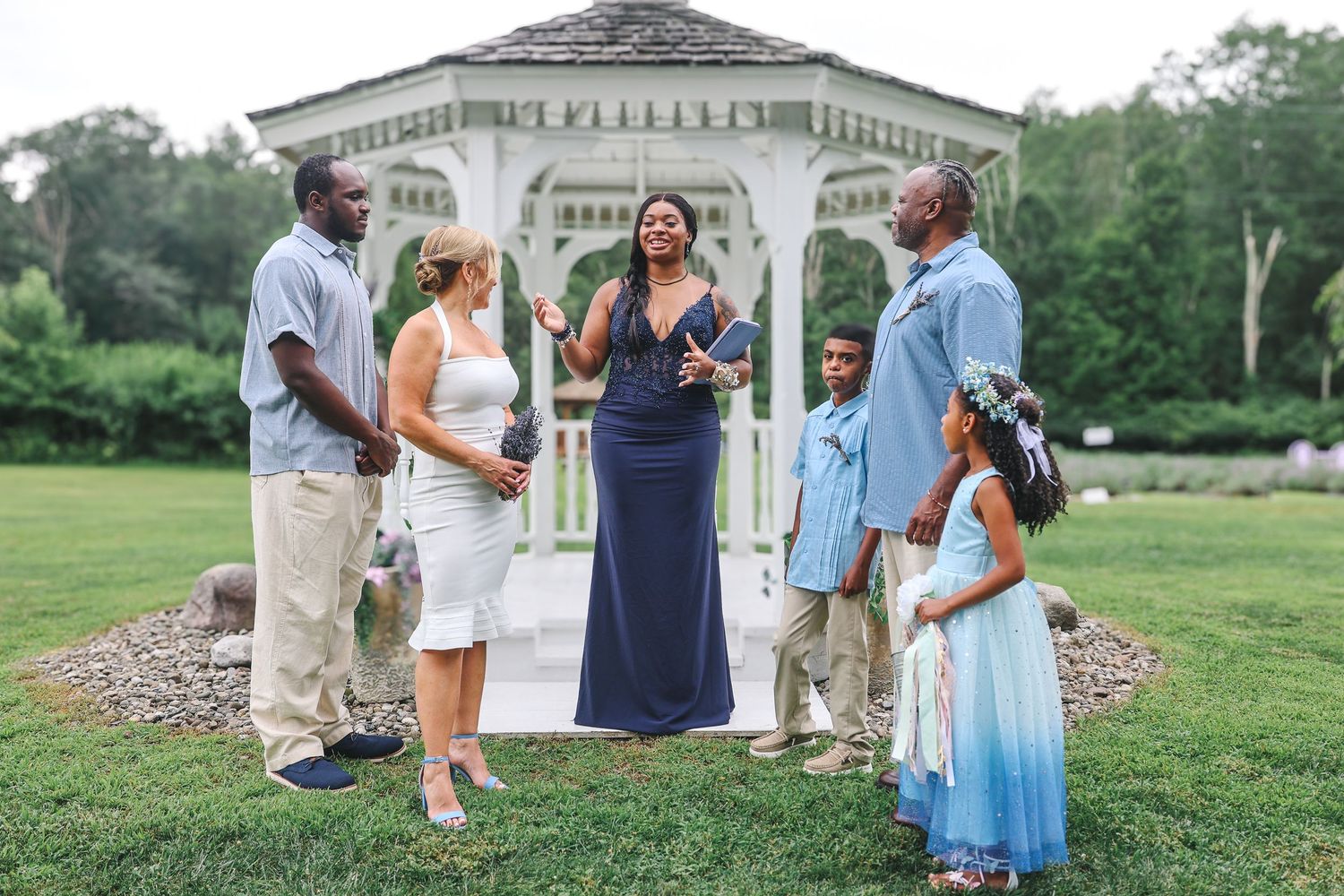 A group celebrates a wedding ceremony outdoors near a white gazebo in a garden setting.