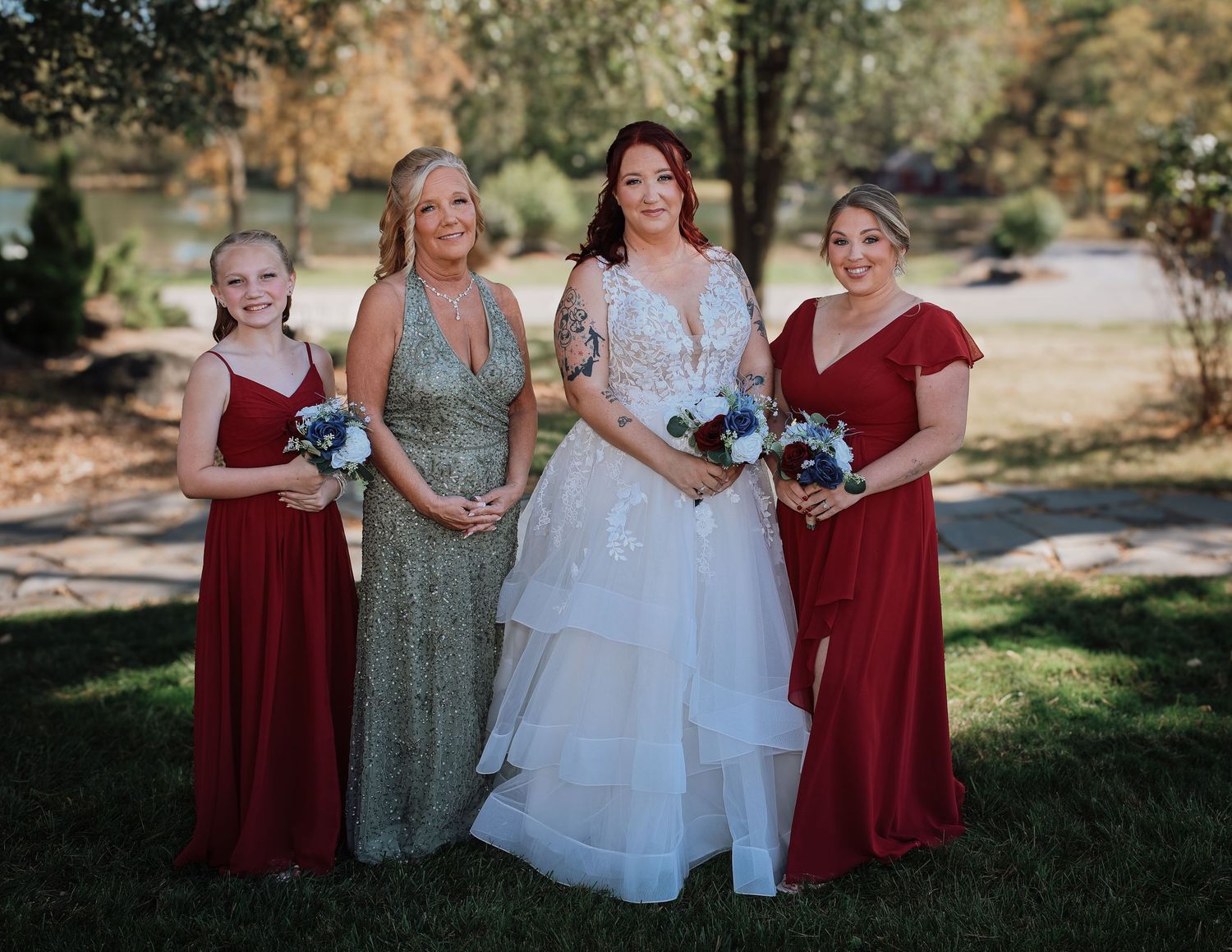 Wedding party members in burgundy and sage dresses pose together on a sunny outdoor lawn.