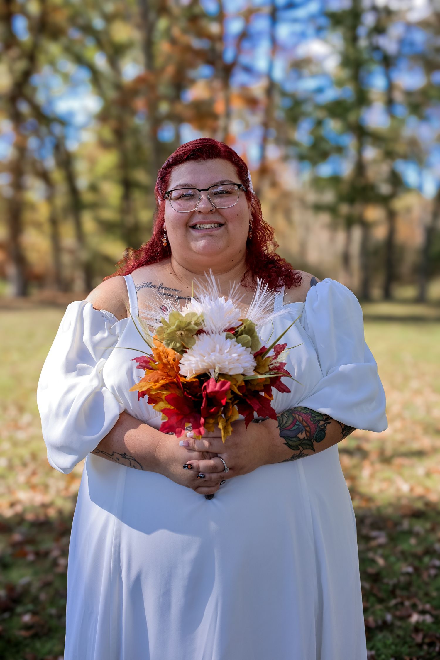 A person in a white dress holds a vibrant autumn bouquet with orange, red and white flowers against a fall landscape.