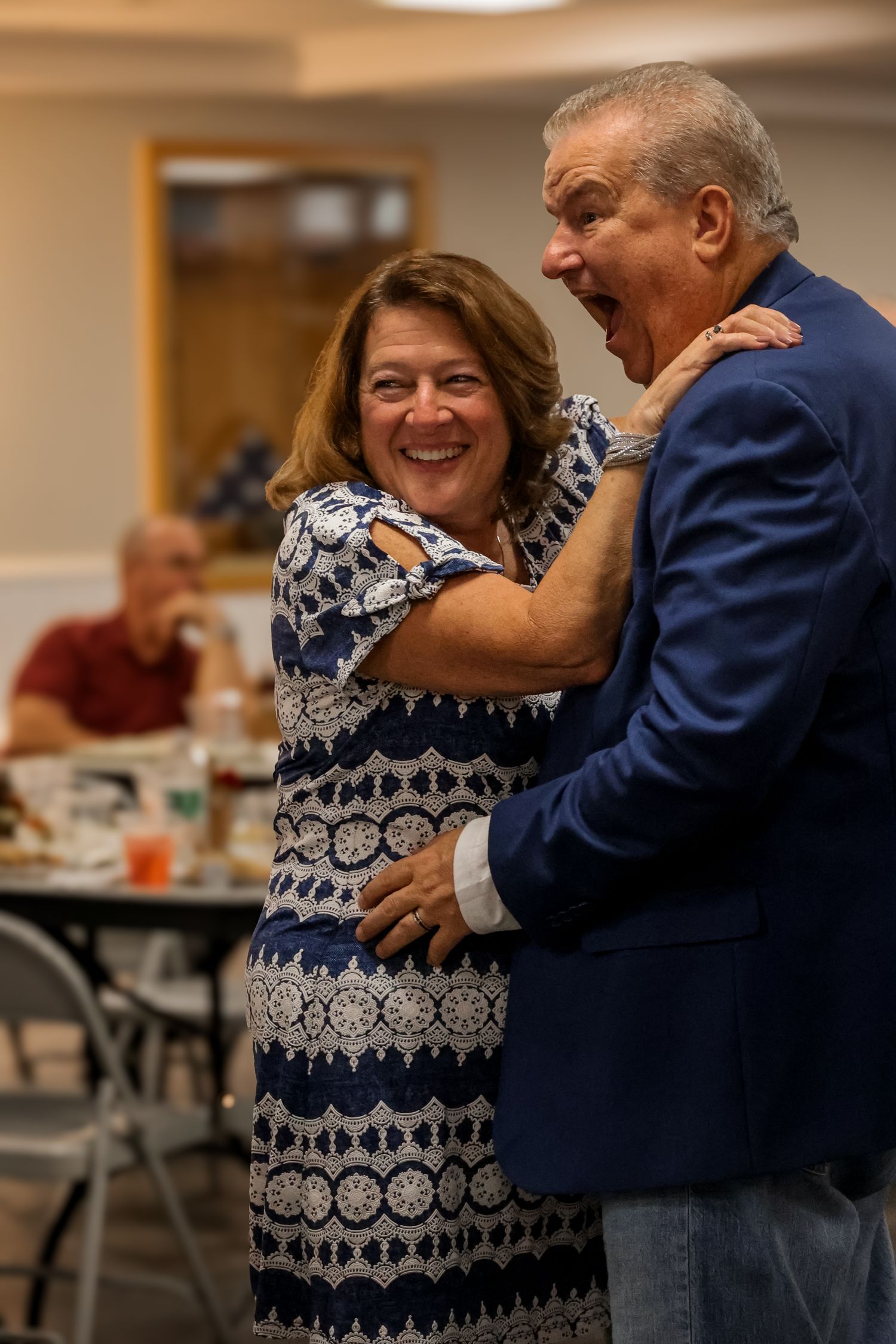 A couple in casual attire share a joyful dance together in what appears to be a community center or social hall.