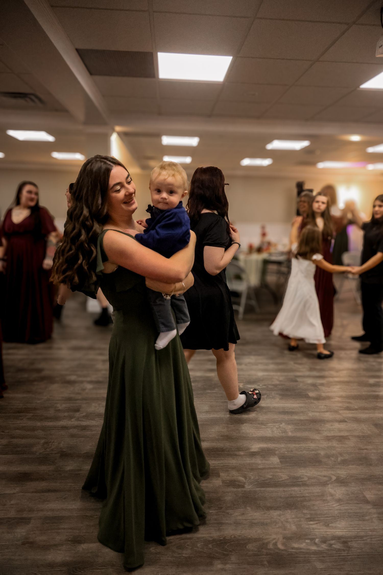 A person in a dark green dress dances while holding a small child in formal attire.