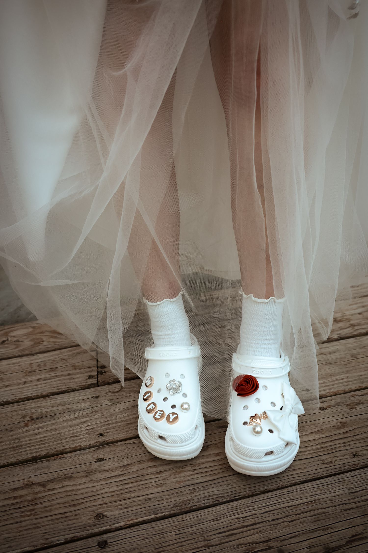 A person wearing a tulle dress and white decorated Crocs shoes stands on a wooden deck.