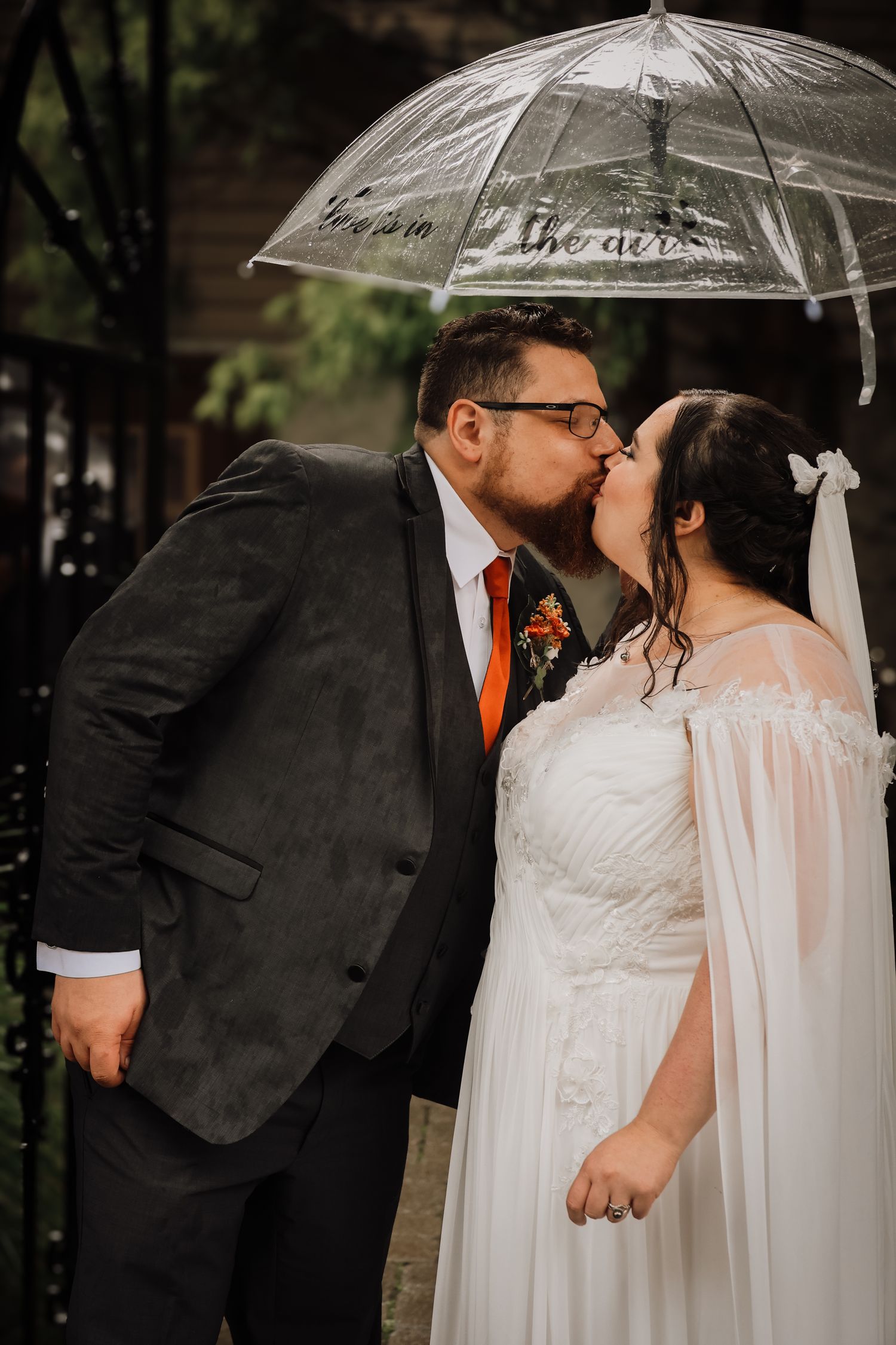 A romantic wedding couple shares a kiss under a clear umbrella on a rainy day.