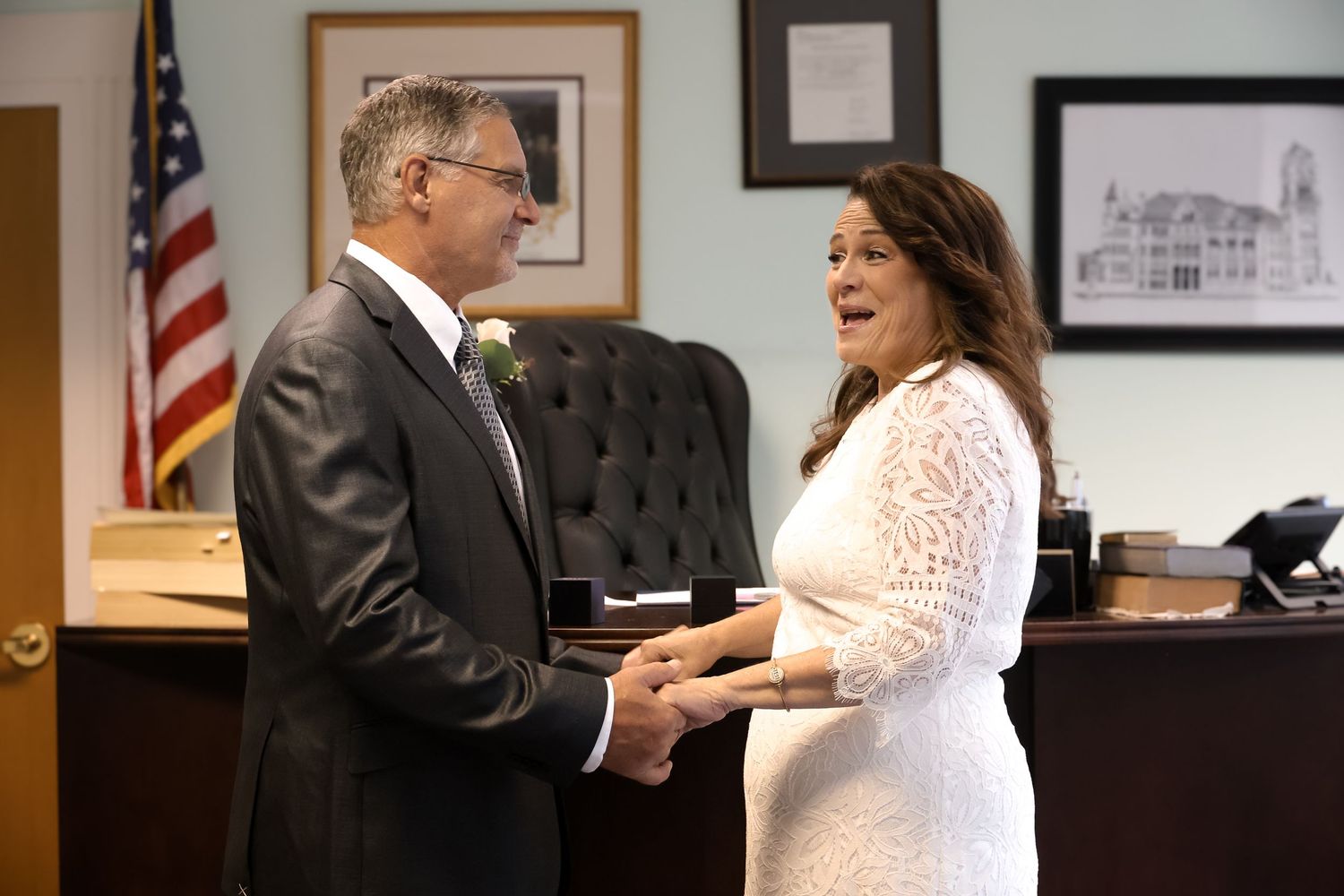 A couple stands together in formal attire during a ceremony in an office with an American flag and framed artwork.