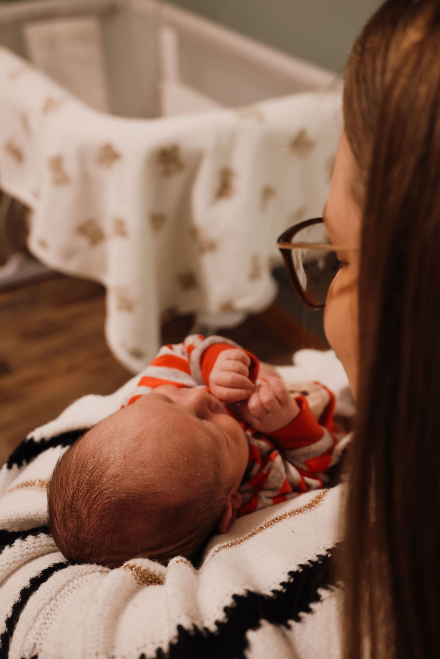 A newborn baby wearing red clothing rests on a black and white patterned blanket while someone in glasses looks down.