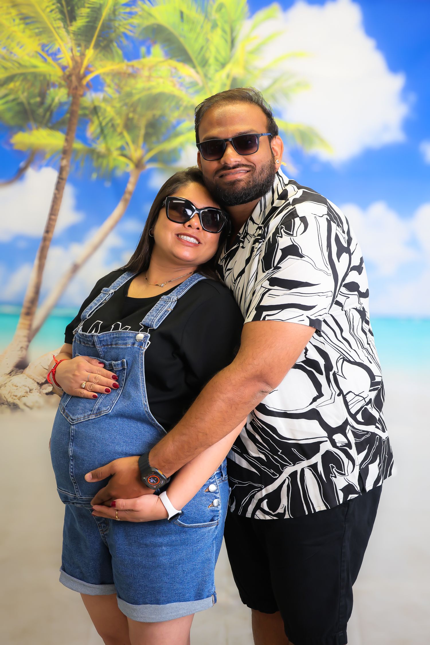 A couple in sunglasses poses together against a tropical beach backdrop with palm trees and blue sky.