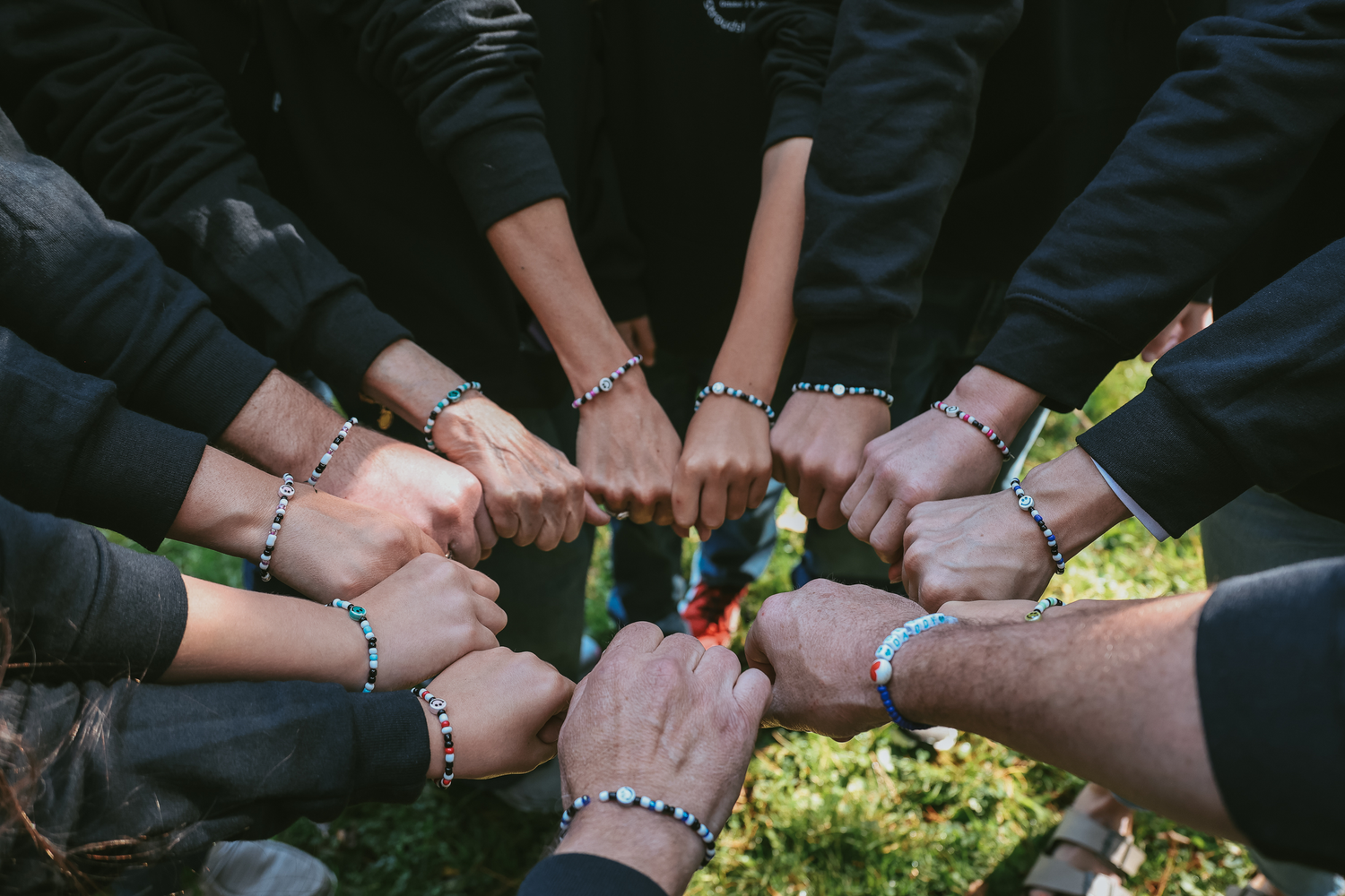 Multiple hands wearing black sleeves join together in a circle formation against a sunlit grassy background.