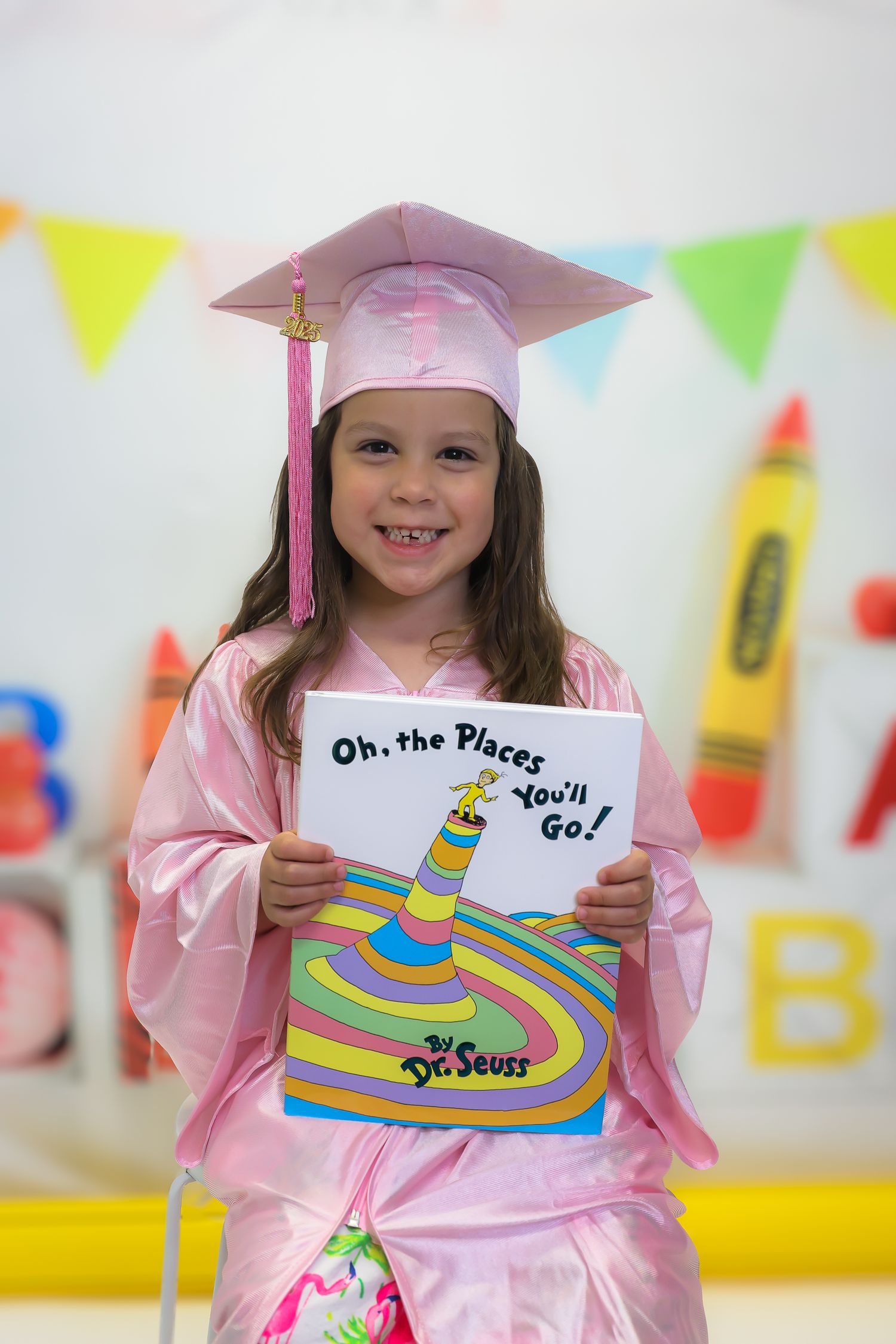 Young girl in pink graduation cap and gown holding Dr. Seuss Oh the Places Youll Go book at kindergarten graduation.