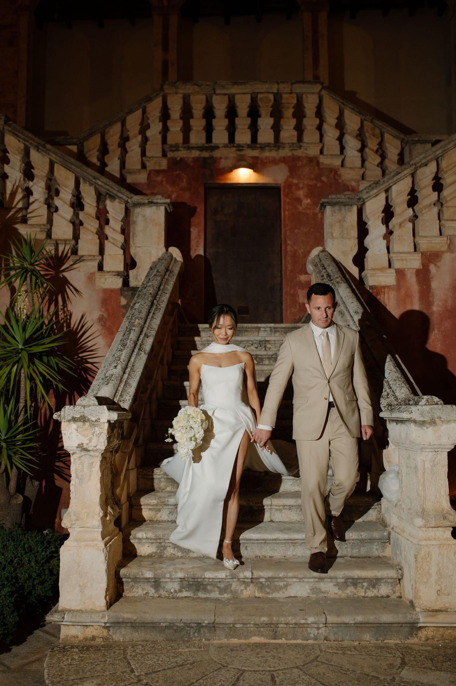A couple descends stone stairs at dusk in formal wedding attire at a historic Mediterranean venue.