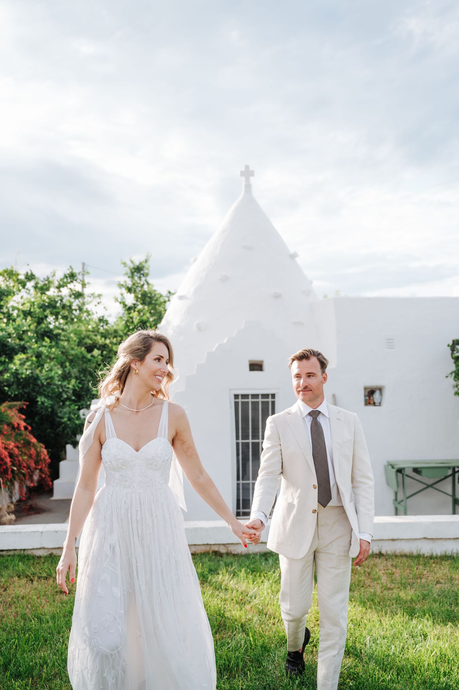 Bride in flowing white dress and groom in light suit walking hand in hand by a white church.