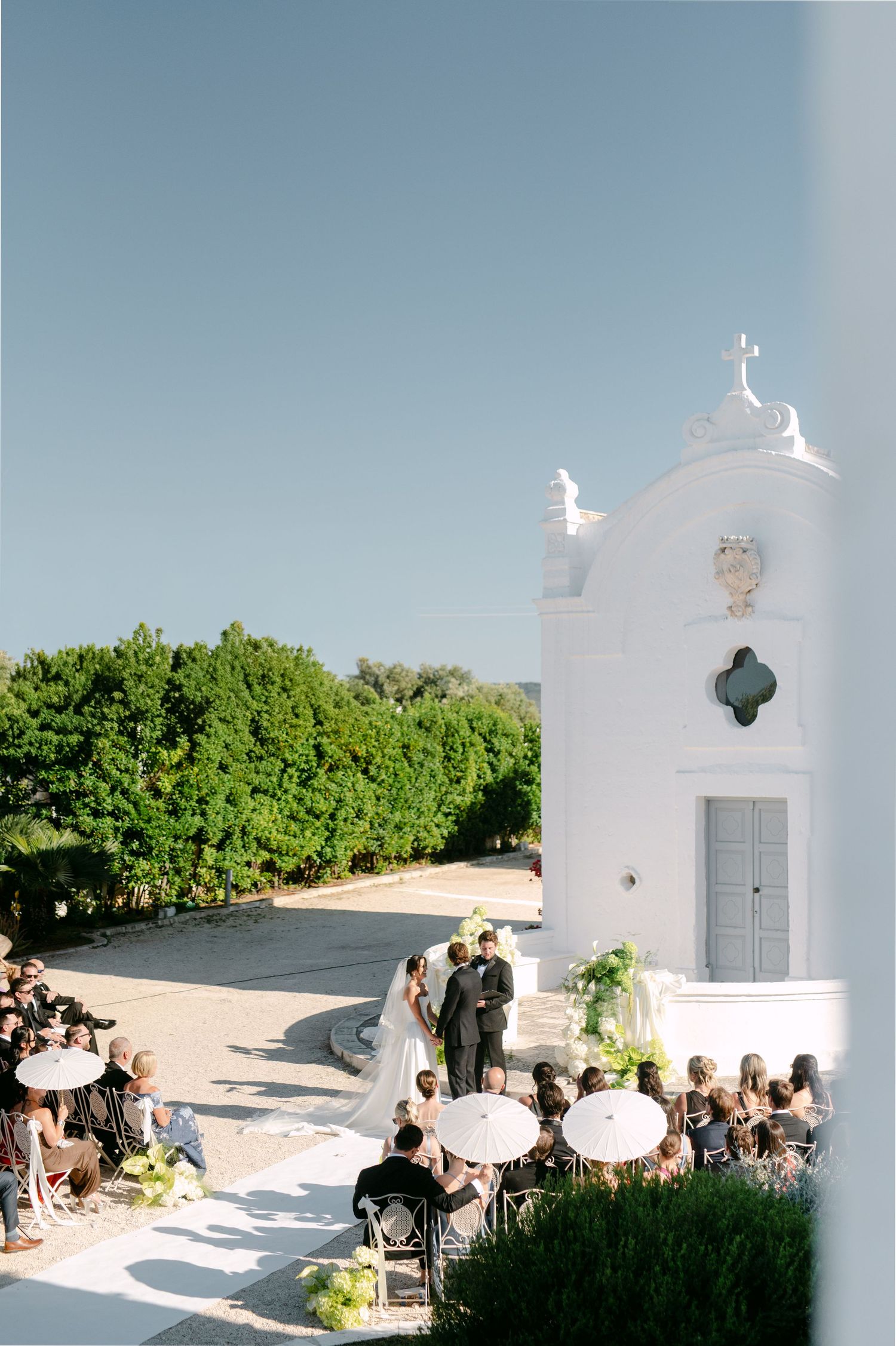 Outdoor wedding ceremony beside a white chapel with guests seated under umbrellas, surrounded by lush greenery under a clear blue sky.