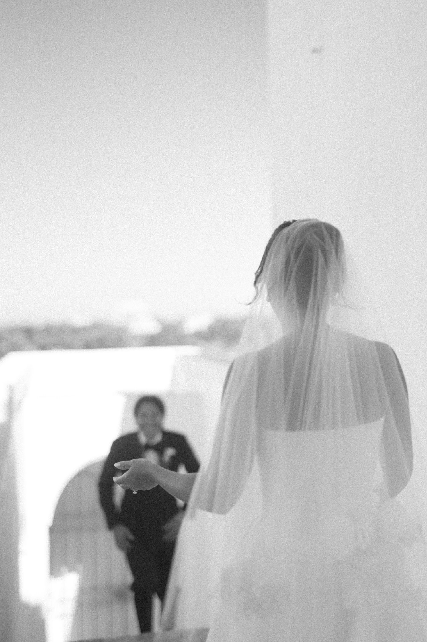 Black and white photo of a bride in a veil walking toward a groom in a tuxedo at an outdoor wedding ceremony.
