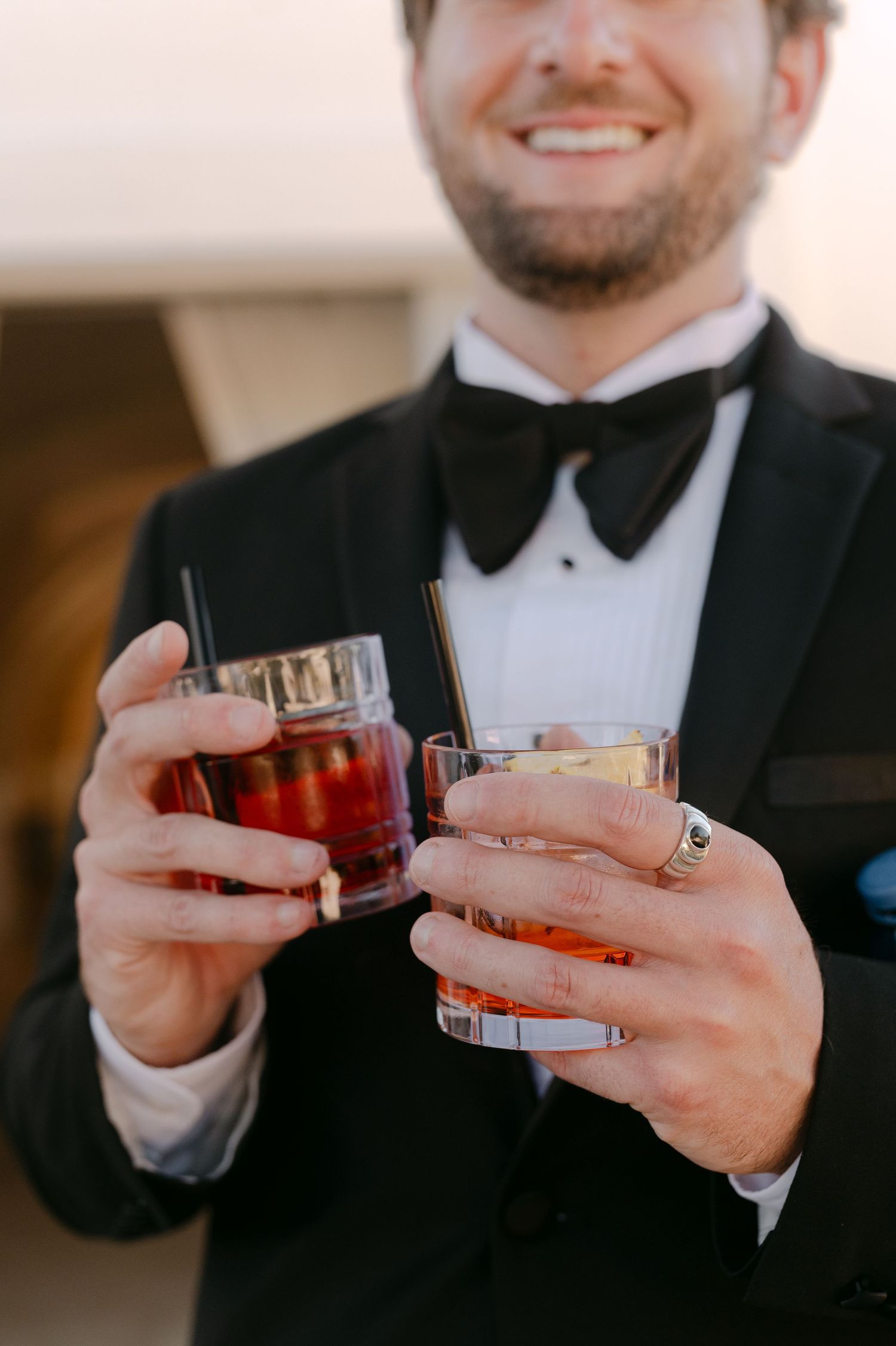 Smiling groom in black tuxedo and bow tie holding two amber cocktails with straws at a formal event.