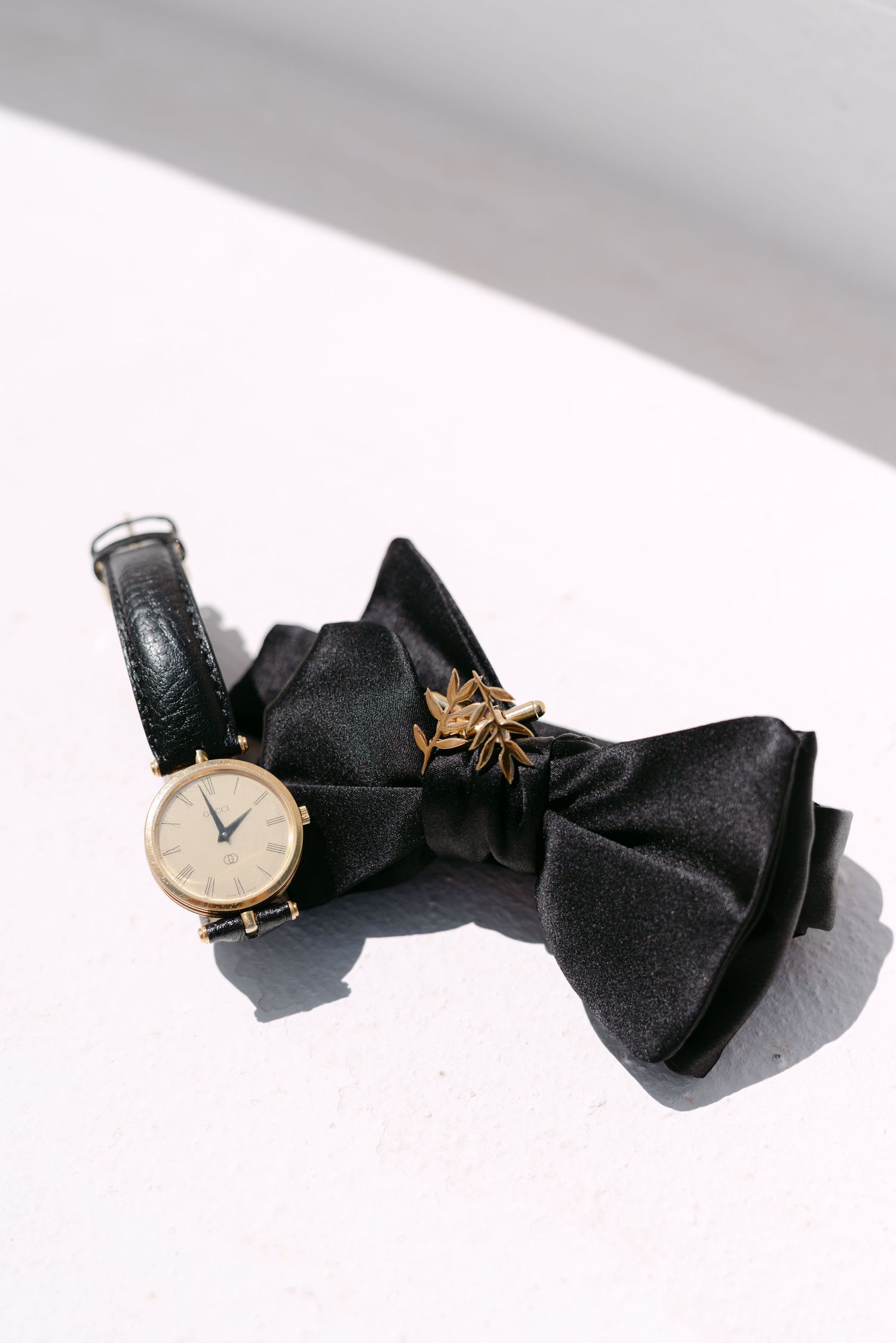 Black bow tie, gold watch with leather strap, and floral lapel pin arranged on white surface.