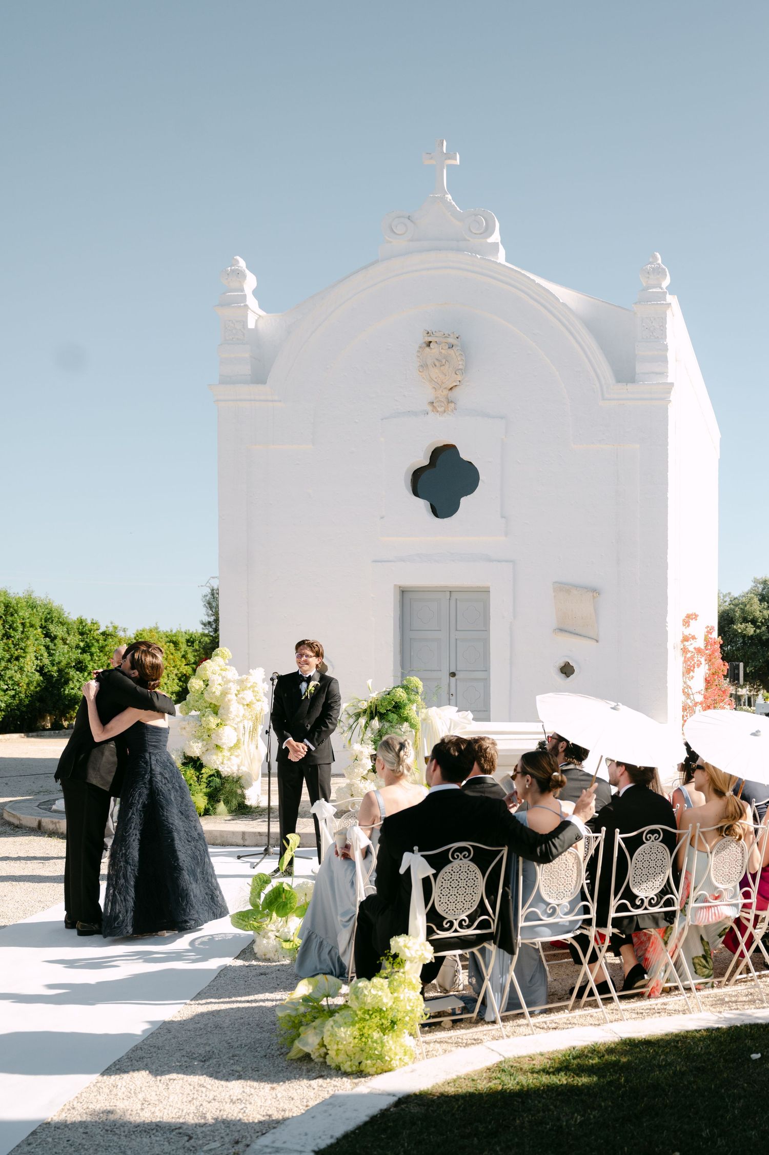 Outdoor wedding ceremony in front of a white chapel, guests seated in ornate chairs with white umbrellas.
