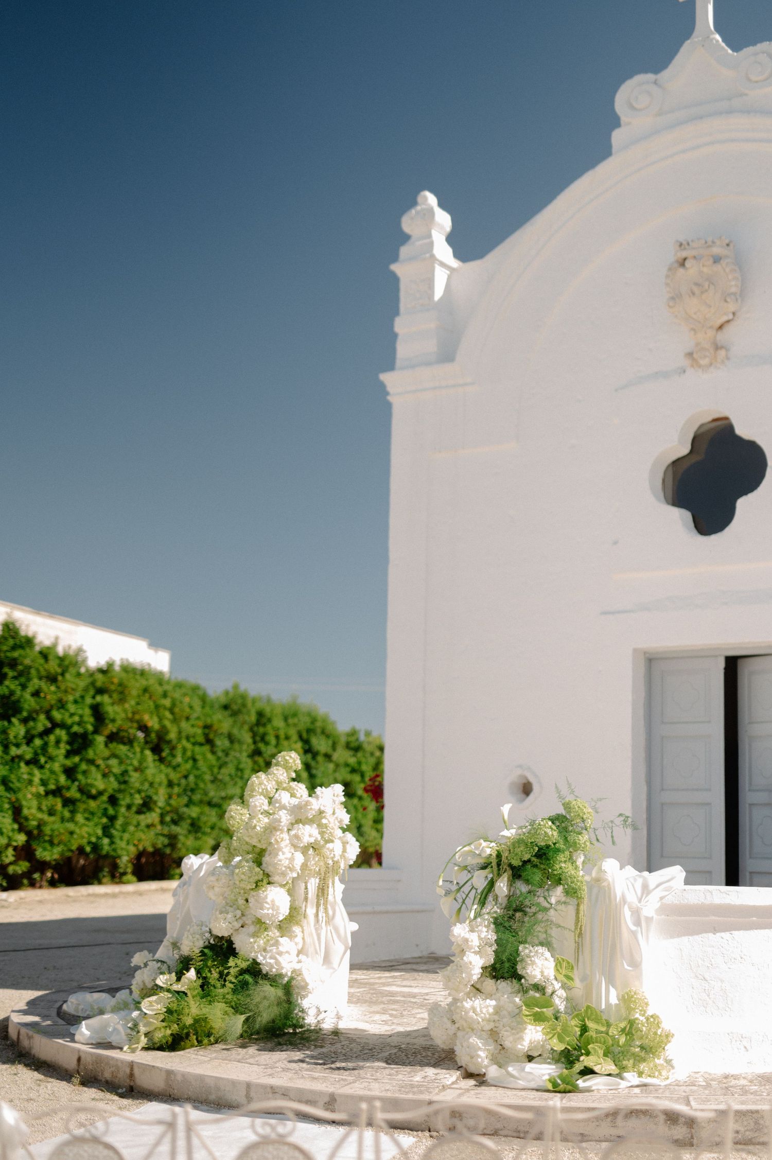 White chapel entrance adorned with elegant white floral arrangements and greenery under a clear blue sky.