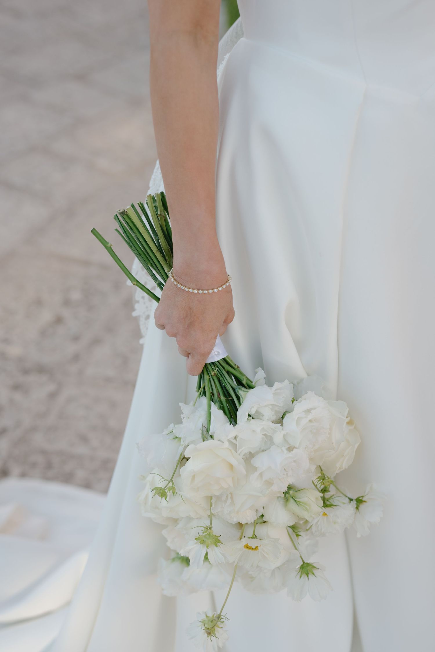 A bride in a white wedding dress holds an elegant bouquet of white flowers against a light stone background.