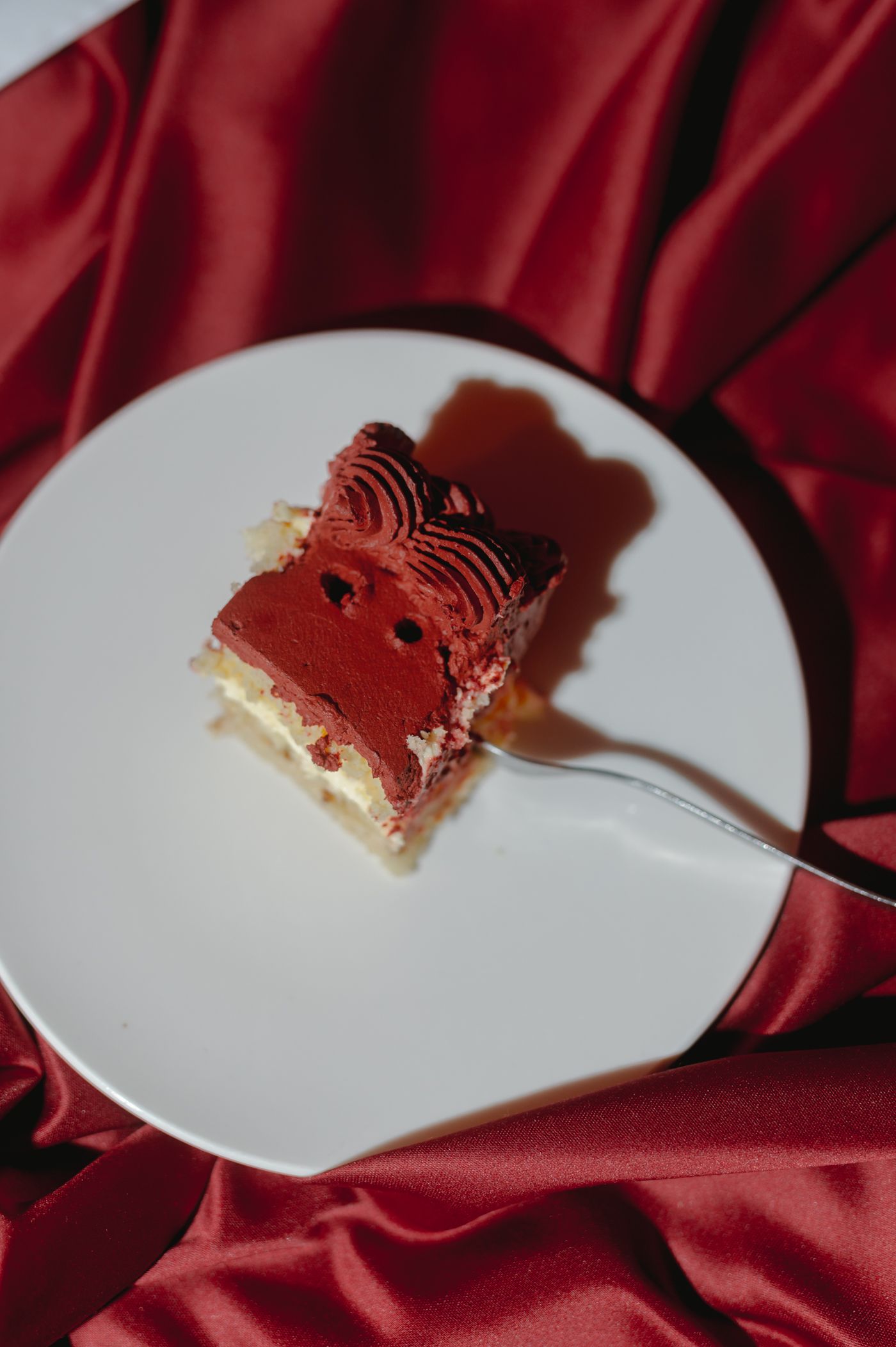 A slice of red velvet cake with layers of cream cheese frosting sits on a white plate against red satin fabric.