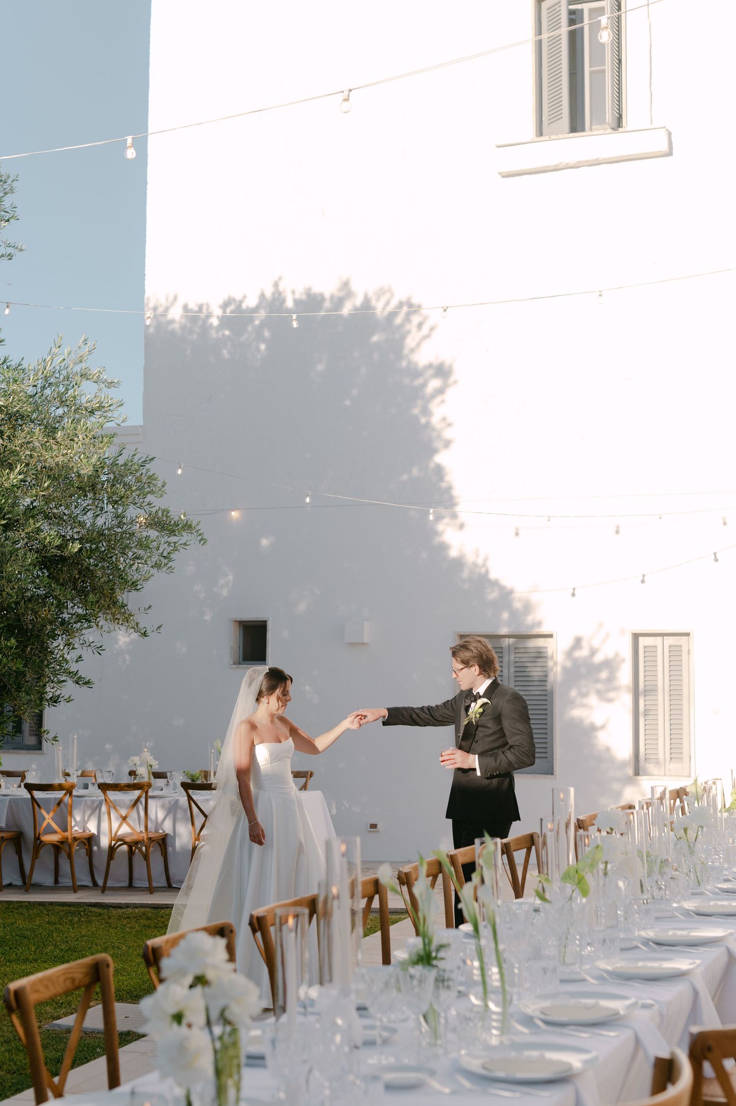 Bride and groom hold hands at elegant outdoor wedding reception with long dining tables set in white venue.