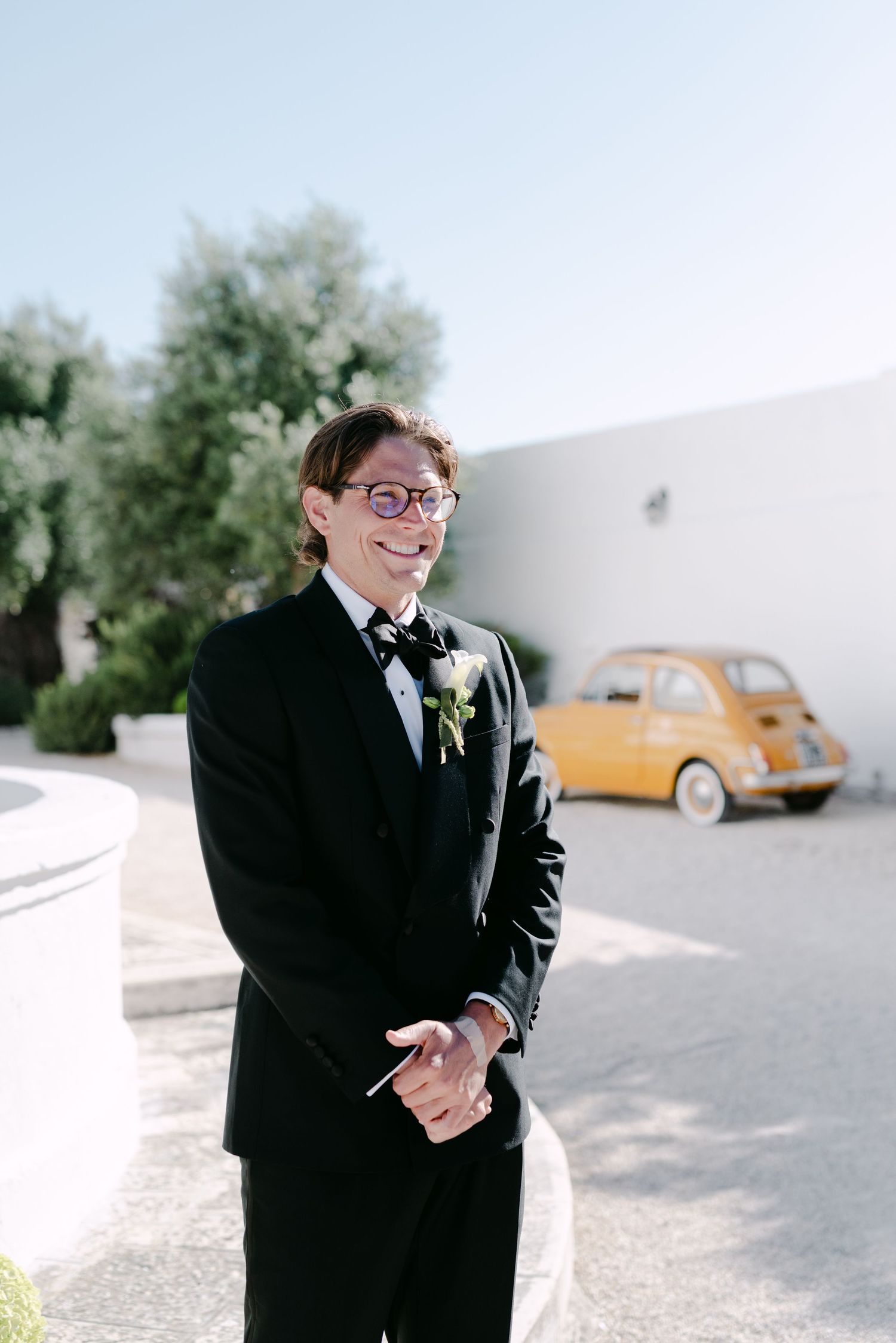 Smiling groom in black tuxedo with boutonniere stands outdoors near vintage yellow Fiat 500 car.