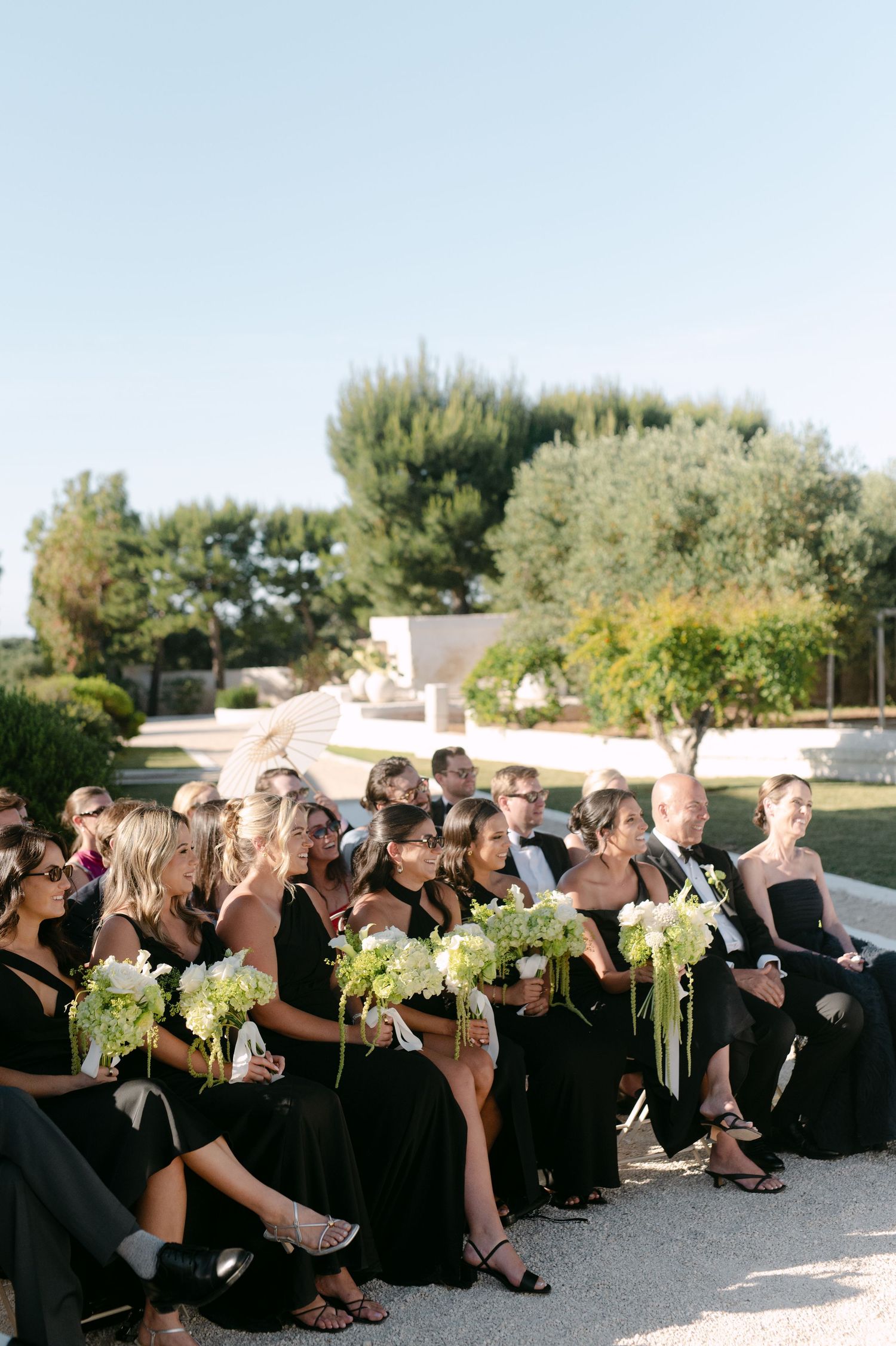 Wedding guests in black attire holding green floral bouquets seated outdoors at an elegant garden ceremony.