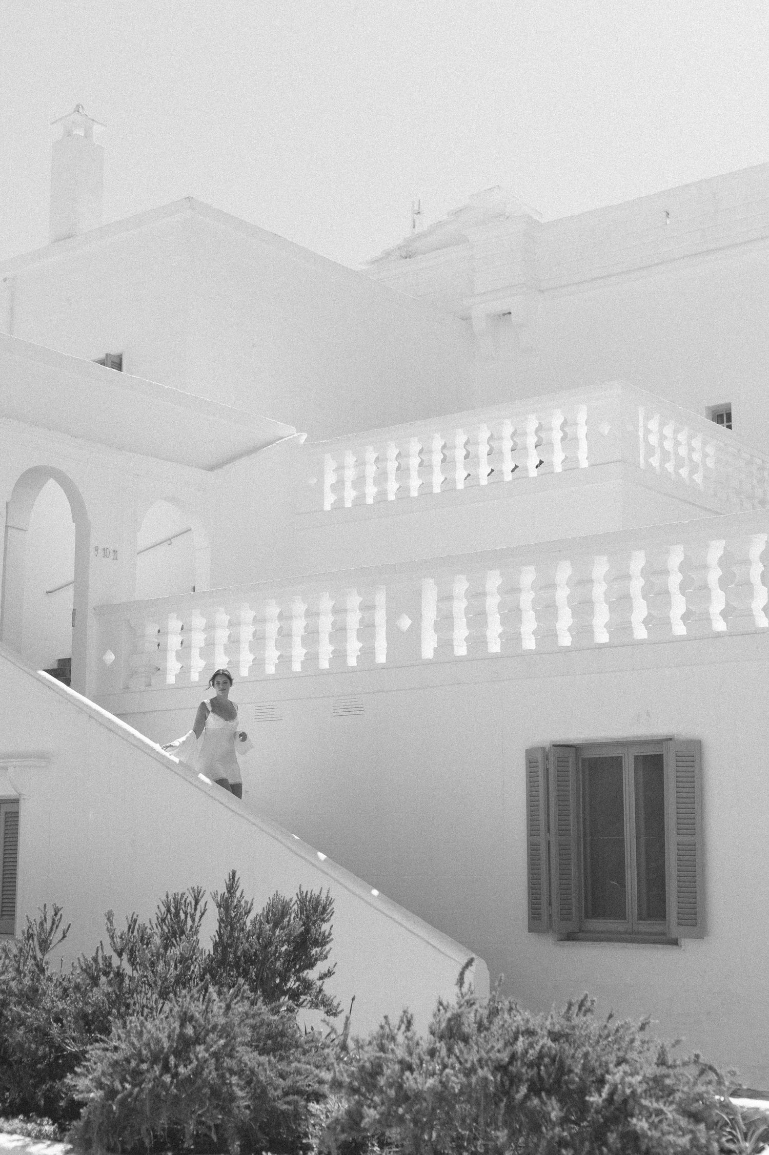 Black and white photo of a person descending white stairs of a Mediterranean-style building with balcony and shuttered windows.