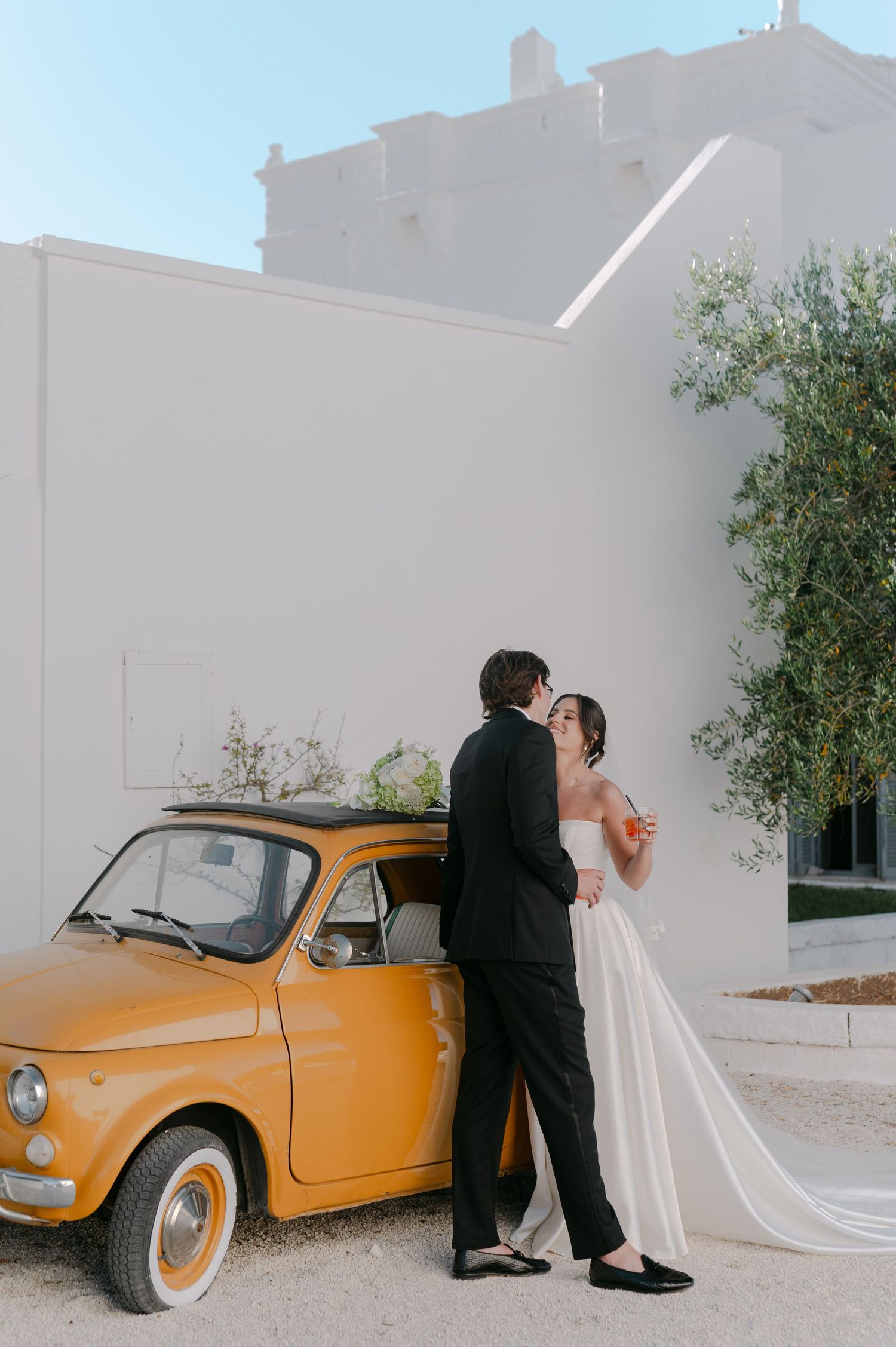 A bride and groom share a kiss leaning against a vintage yellow Fiat 500 outside a white Mediterranean villa.